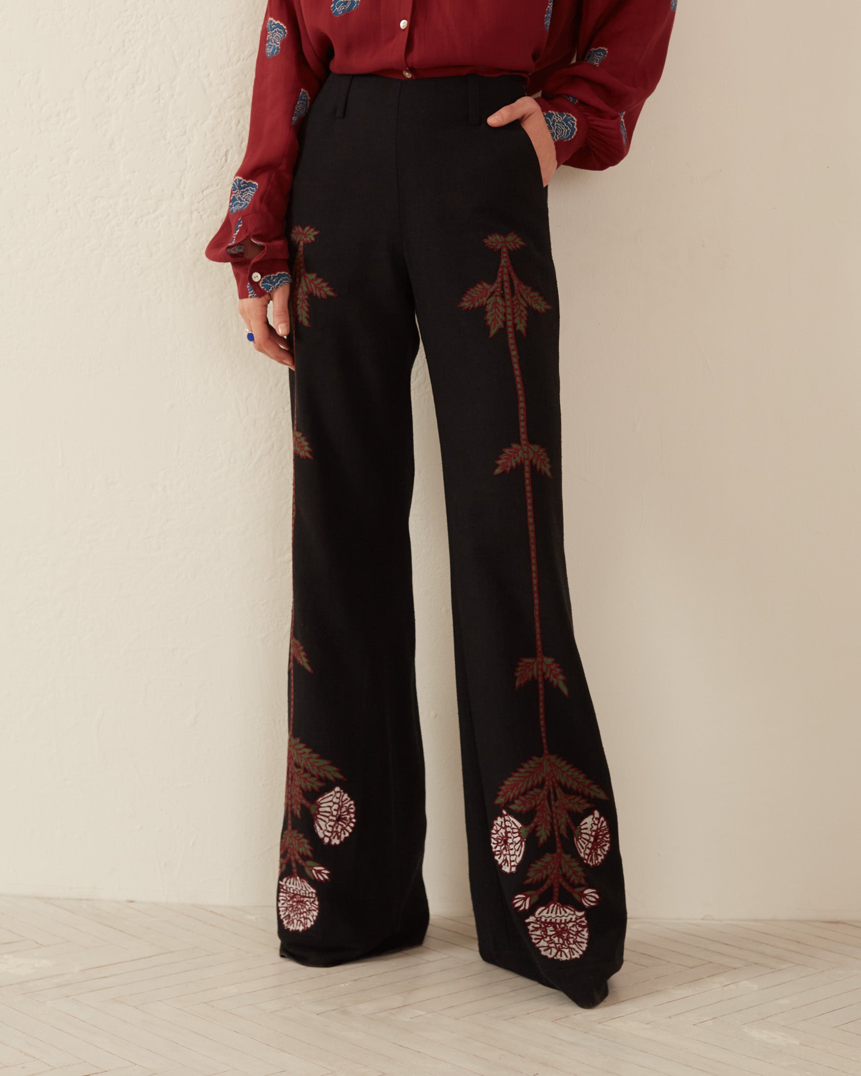 A person wearing the Charlie Black Bloom Embroidered Pants, high-waisted and adorned with intricate floral motifs, stands against a light-colored wall. Made from elegant Indian silk slub, these pants feature large floral designs on the legs and are paired with a long-sleeve burgundy blouse.