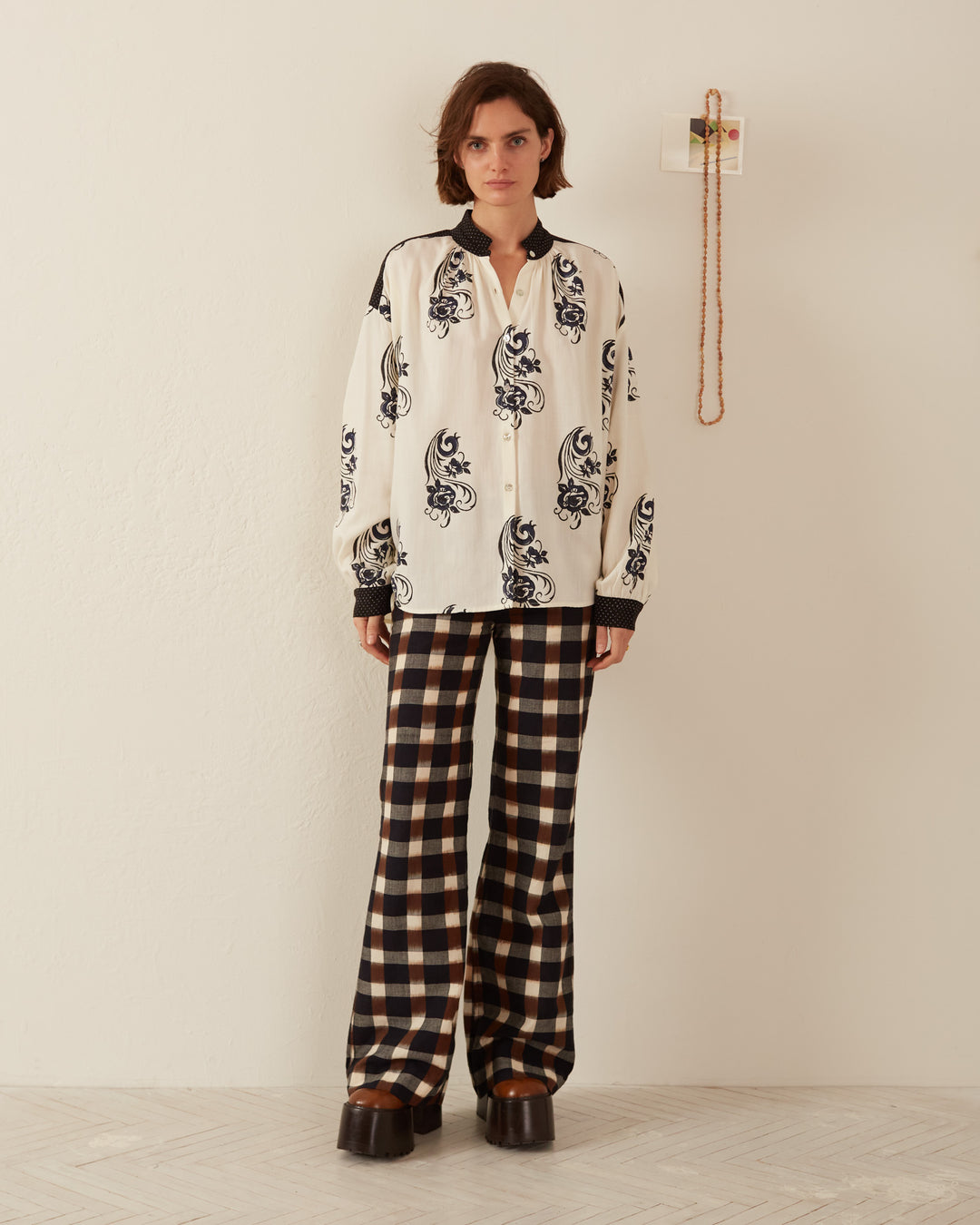 A person stands against a light-colored wall, dressed in a white Kiki Noir Shirt embellished with a black floral design and black-and-white checkered wide-leg pants. They have short hair and are wearing brown platform shoes. A string of beads on the wall casts shadows on the textured cotton fabric.