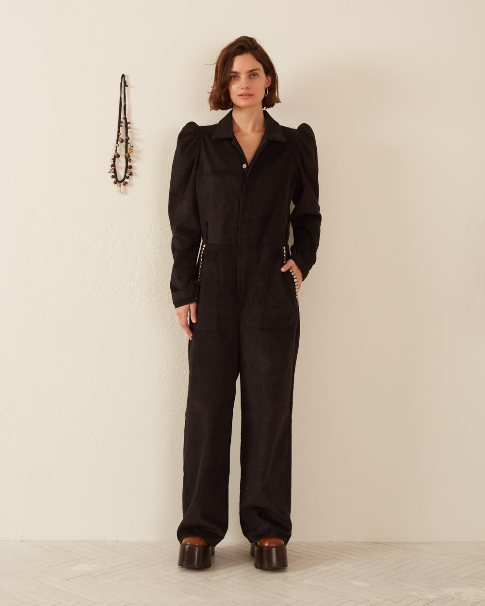 A person wearing the Minnie Onyx Corduroy Jumpsuit with stud details and large platform boots stands against a plain white wall, with a necklace hanging beside them.