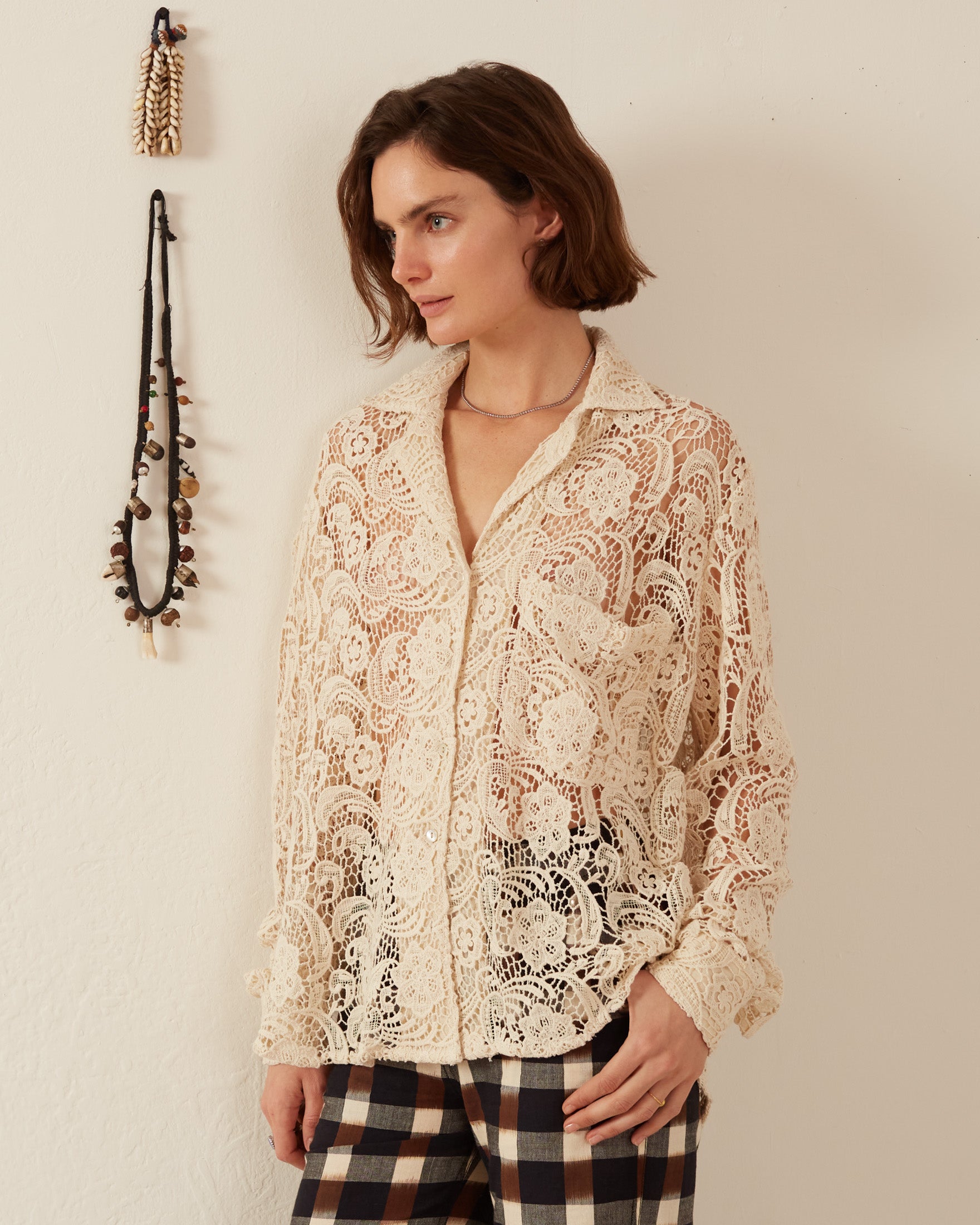 A person wearing the Patti Ivory Floral Lace Shirt with a relaxed fit and plaid pants stands against a white wall. A decorative string with small items hangs on the wall. The v-neck shirt complements their neutral expression as they gaze to the left.