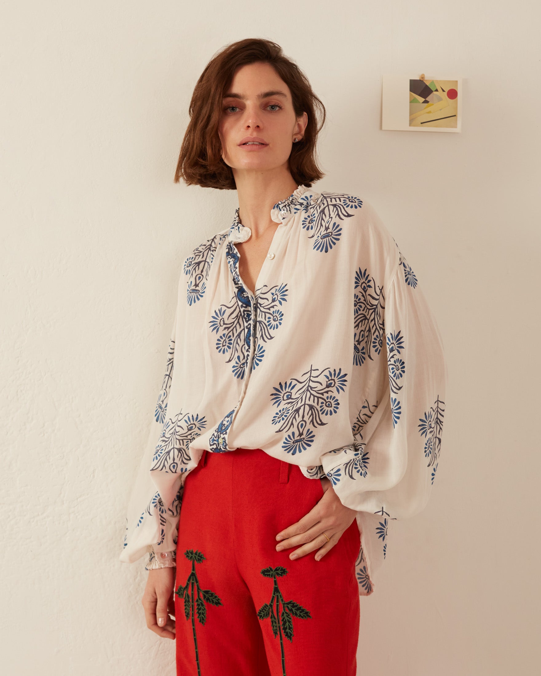 A person stands against a cream-colored wall, wearing a Poet Black and Blue Blouse with intricate floral patterns, paired with red pants adorned with floral embroidery. The ruffled collar adds charm, while an abstract painting hung nearby complements the casual, artistic style.