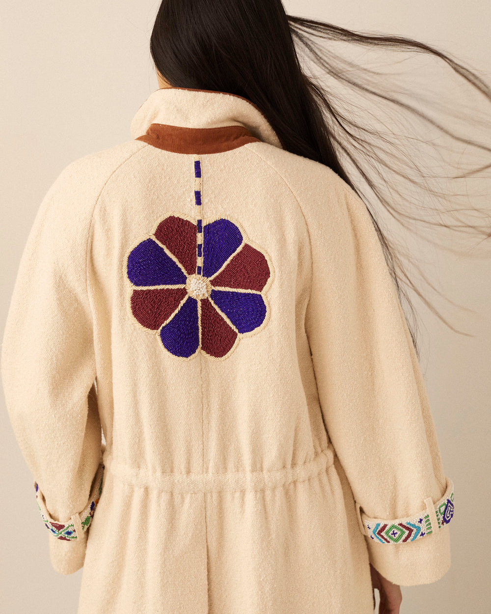 A person with long hair, shown from behind, wears the sophisticated Wren Harvest Trench Coat in cream. It showcases a large, embroidered flower on the back and patterned cuffs with brown accents. The design incorporates purple and maroon tones, complemented by a hand-beaded collar for an exquisite touch.