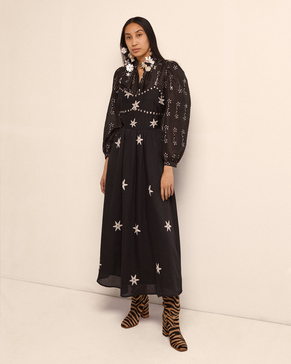 A person stands against a plain backdrop, wearing the Autumn Western Star Dress, featuring black fabric adorned with white star and geometric patterns, crafted using traditional methods. They have long hair and are accessorized with large star-shaped earrings and tiger-striped boots.