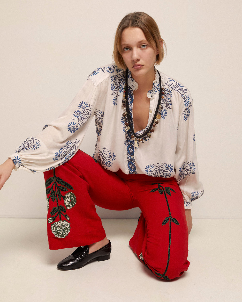 Wearing a white blouse adorned with blue embroidery, a person gracefully kneels in the exquisite Charlie Red Bloom Pant, which features intricate floral motifs. With long hair cascading down their back, they gaze intently at the camera against a plain backdrop, embodying the elegance of hand-embroidered artistry.