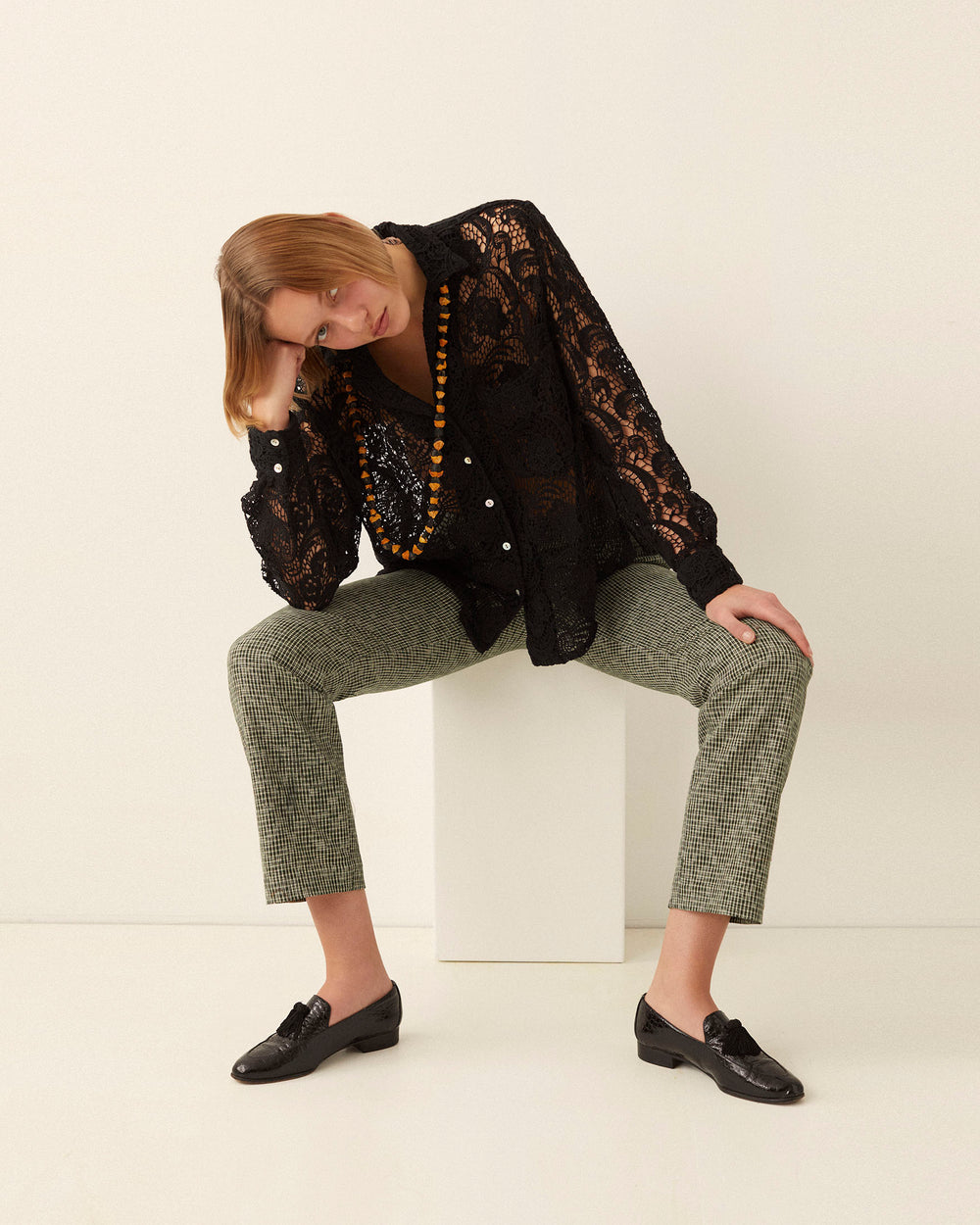 A person with chin-length hair sits on a white cube, leaning forward with one hand on their head. They wear the Patti Black Floral Lace Shirt, which features a relaxed fit, along with gray plaid pants and black loafers. Their look is accessorized with a long orange beaded necklace against a plain white background.