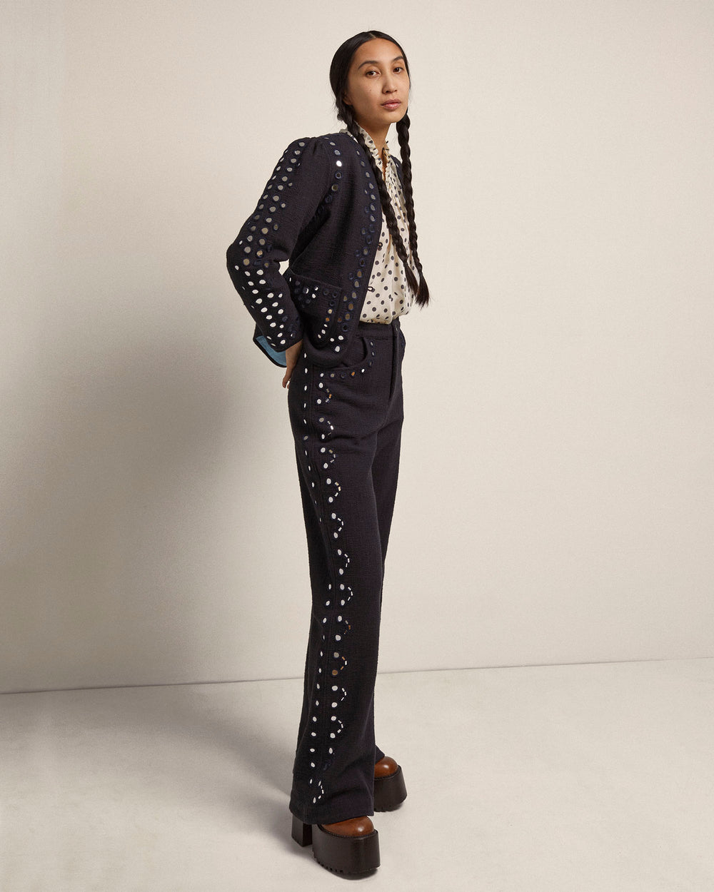 A person with long braided hair stands against a plain background, wearing the Billie Midnight Mirror Jacket paired with a dark pantsuit. The outfit is complemented by a patterned shirt and platform shoes, highlighting the elegance of traditional methods.