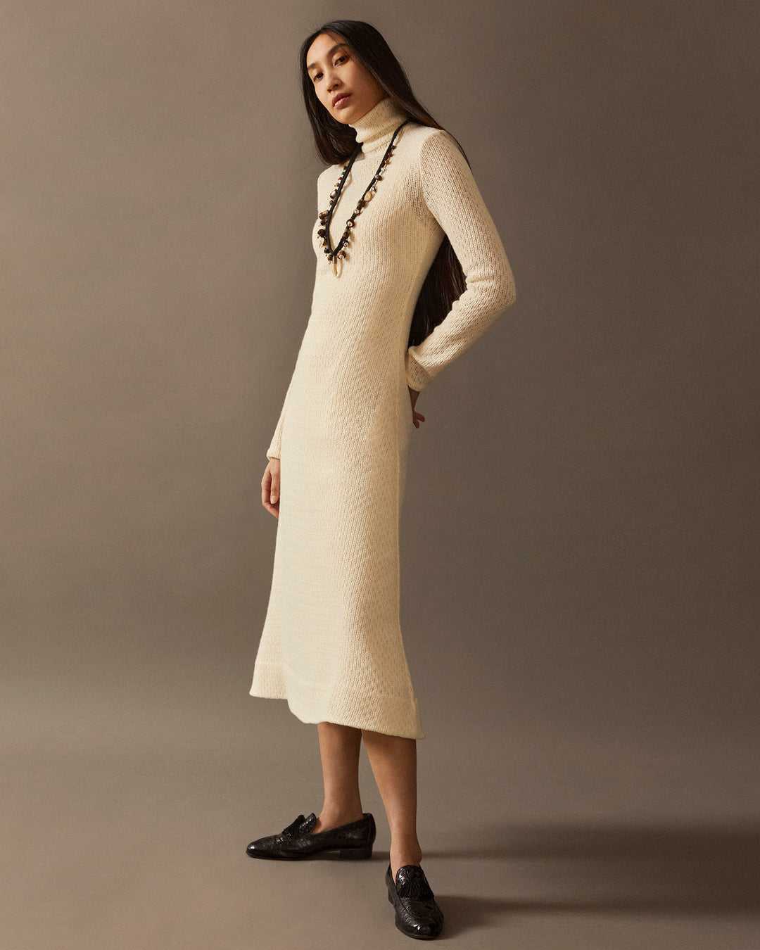 Clad in the elegant Nona Marble Knit Dress, a person with long hair showcases the cream-colored, long-sleeve Italian wool turtleneck against a plain backdrop. Pairing it with black shoes, they strike a pose with one hand on their hip, exuding timeless charm.