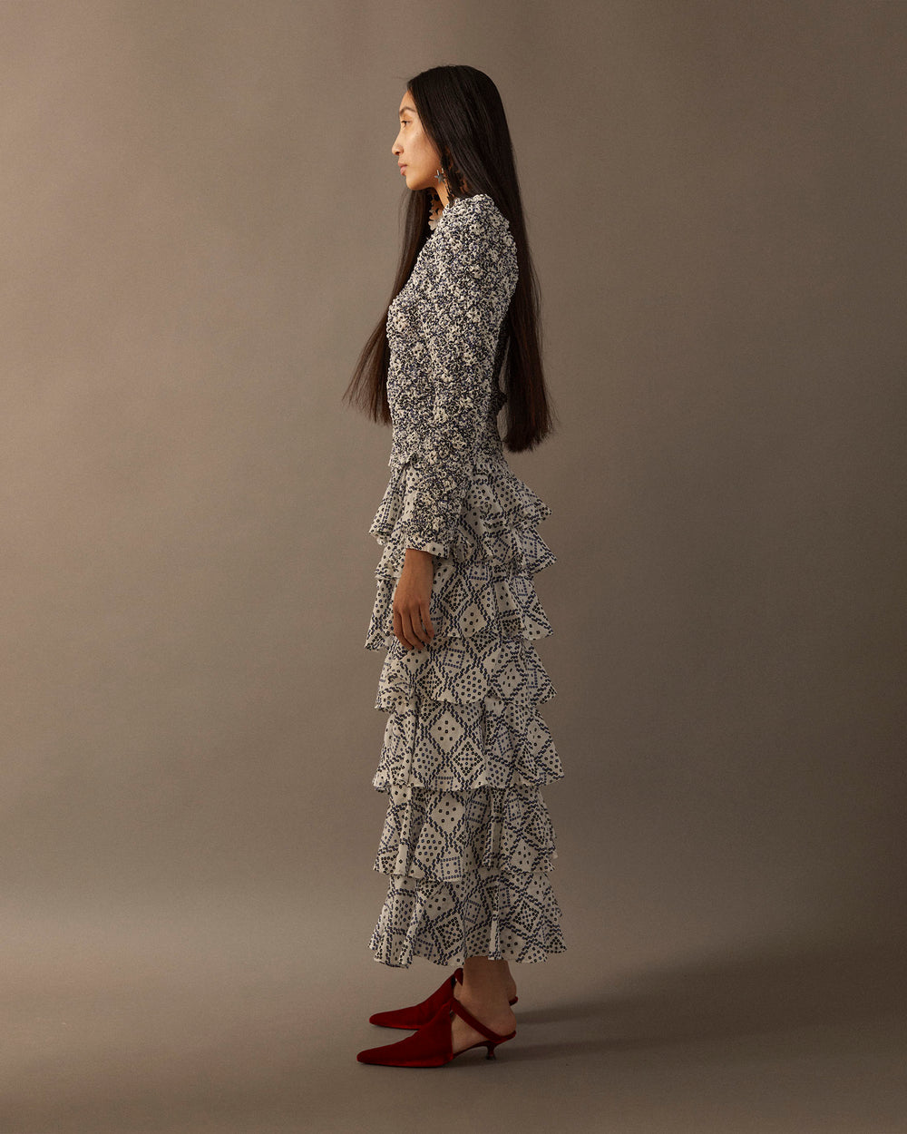 A woman with long dark hair stands sideways against a neutral background. She is dressed in a hand-printed Tencel ensemble, showcasing the Maisie Domino Smocked Top with multiple ruffles, paired elegantly with red heeled shoes.