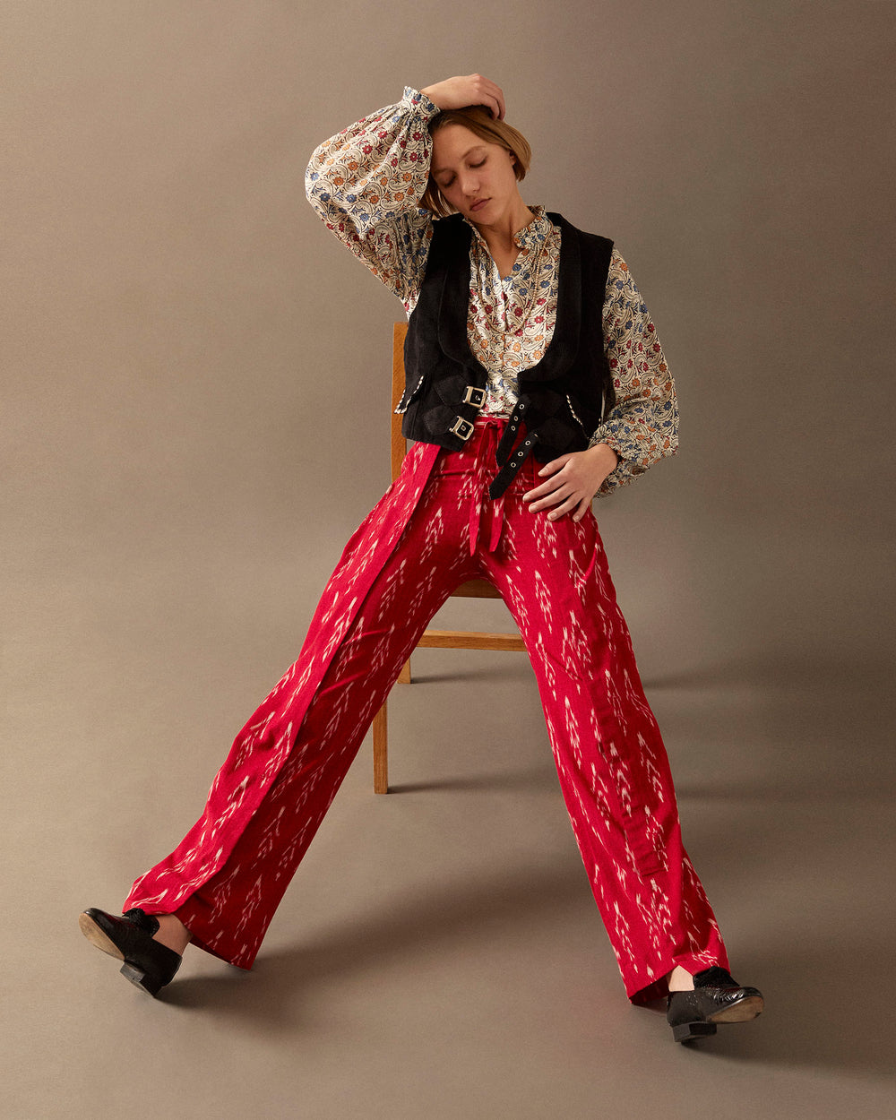 A person sits on a wooden chair against a plain background, wearing a patterned blouse and the Lucien Cotton Vest adorned with silver studs, paired with bright red patterned pants. Their head is tilted, and one arm rests on their knee.