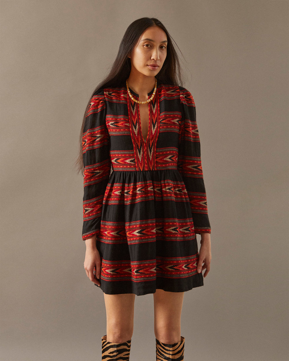 A woman with long dark hair wears a vibrant Winslow Arrow Dress, showcasing black, red, and orange geometric designs. Crafted using traditional methods, the dress complements her gold necklace as she stands against a neutral background.