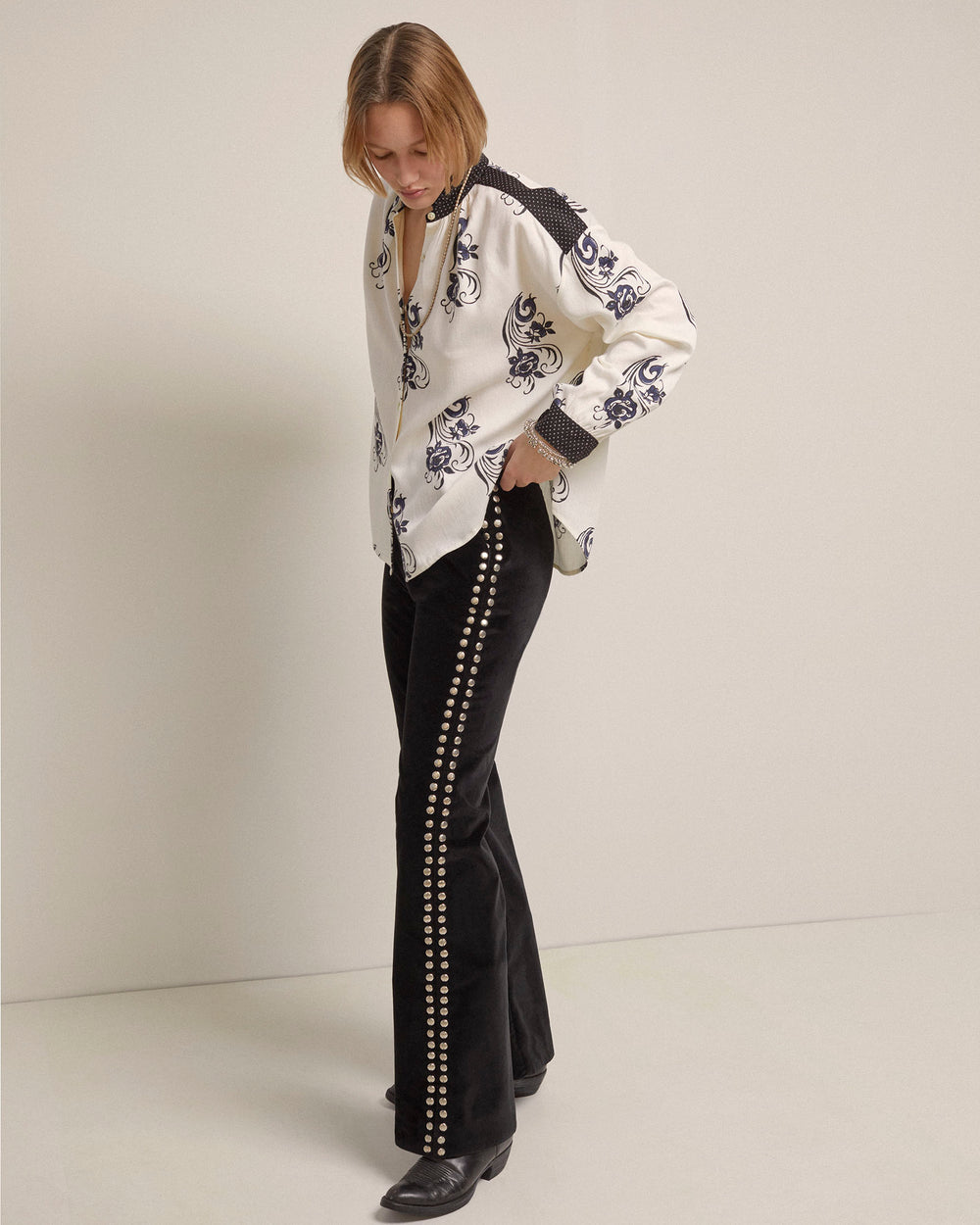 A person is wearing a white embroidered blouse paired with the black Charlie Rodeo Velvet Pant, featuring a line of silver studs along the sides. The pants have a subtle flared leg that adds elegance to the ensemble. They stand against a plain background, looking downward with their hands in pockets, exuding quiet confidence.