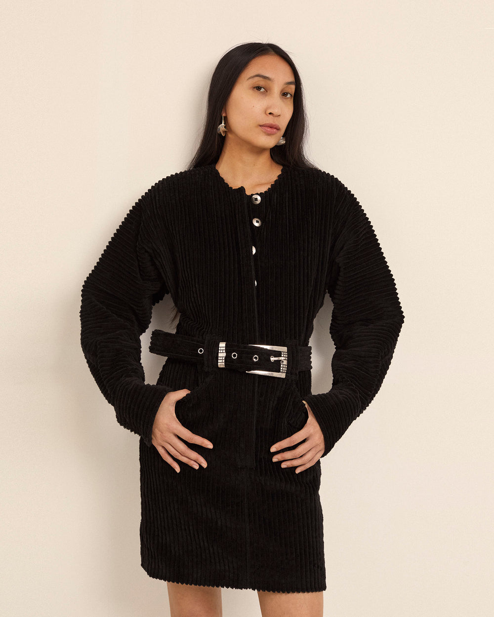 A woman is posed against a simple backdrop, showcasing the sophisticated Laurent Licorice Corduroy Dress, expertly crafted from Italian wide wale corduroy. This black dress boasts long sleeves and an adjustable belt that beautifully emphasizes her waistline. With long dark hair cascading down her shoulders, she exudes confidence as she gazes directly into the camera with her hands on her hips.