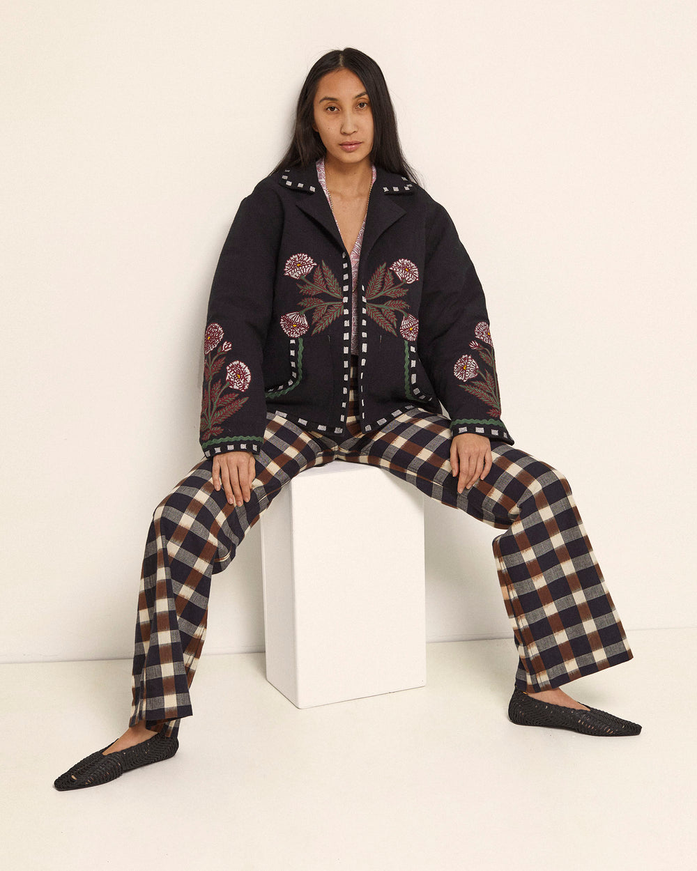 A person is seated on a white cube, wearing a black jacket adorned with floral embroidery and Charlie Plaid Princess Pants. They sport long dark hair and are dressed in black woven shoes. The backdrop of a plain white wall highlights the hand-embellished details of their ensemble.