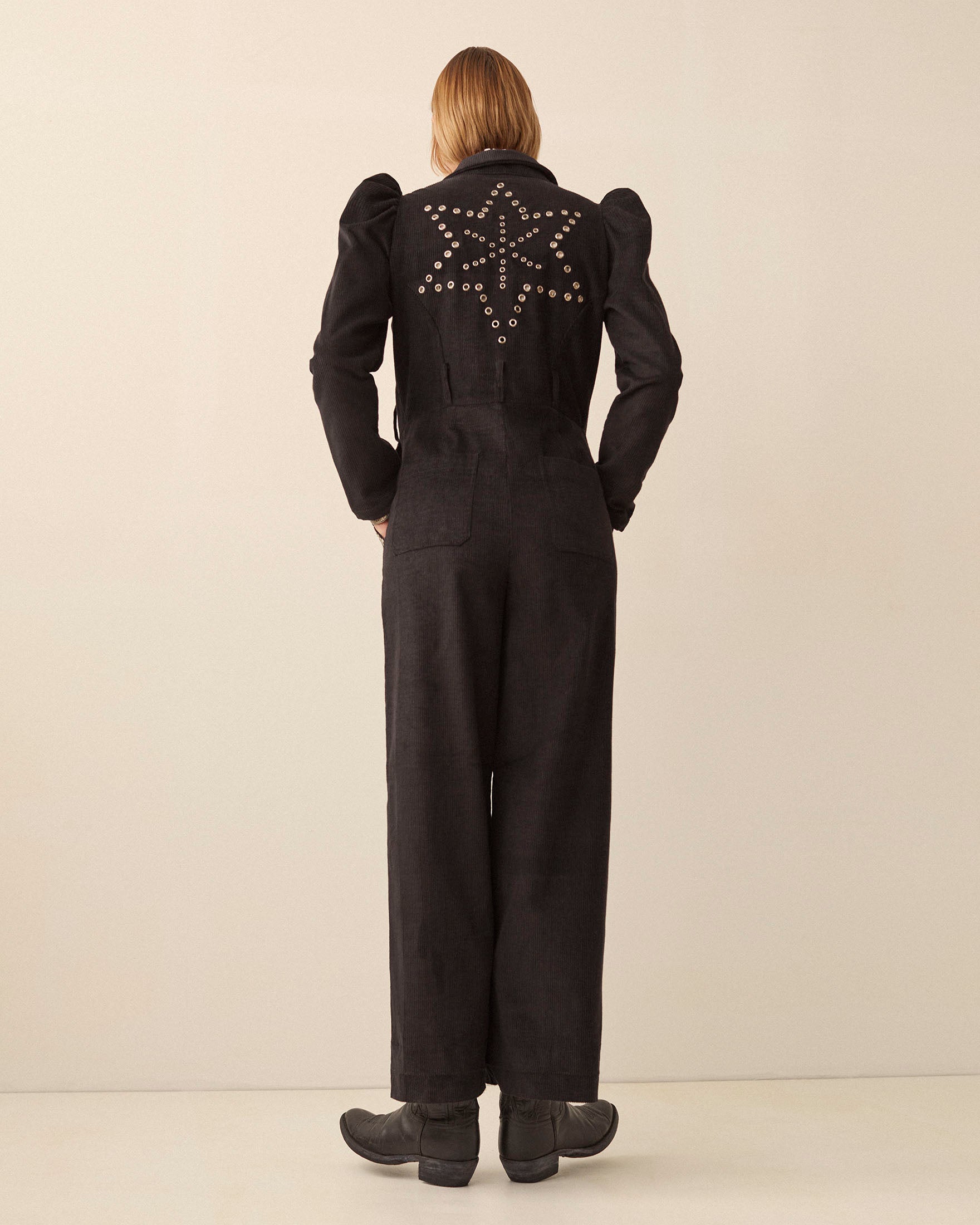 A person wearing the Minnie Onyx Corduroy Jumpsuit with star-shaped stud details stands facing a plain, light-colored wall. Their shoulder-length hair falls neatly, and they pair the look with black shoes. The outfit features puffed sleeves and wide legs for a striking silhouette.