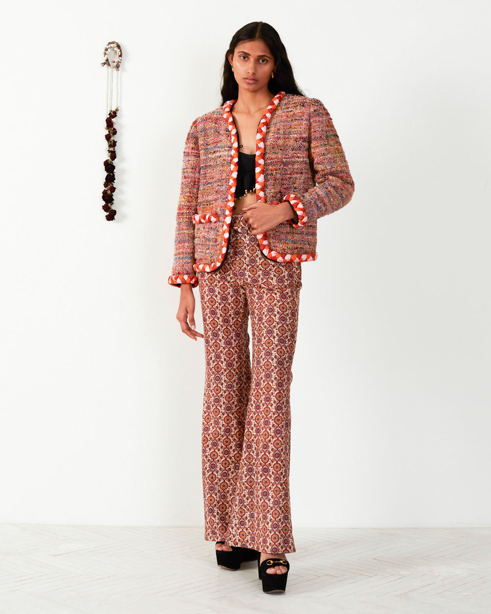 A person in a Billie Rose Dust Boucle Jacket and patterned flared pants made from recycled fibers stands against a plain wall with a decorative hanging.