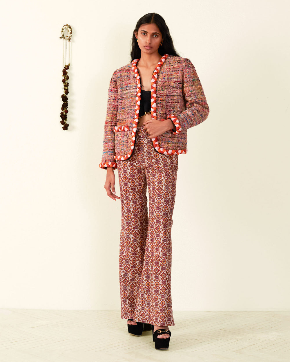 A person stands against a white wall wearing the Billie Rose Dust Boucle Jacket with red and white trim, paired with patterned flare pants and black platform heels made from recycled fibers. A decorative wall hanging is visible in the background.