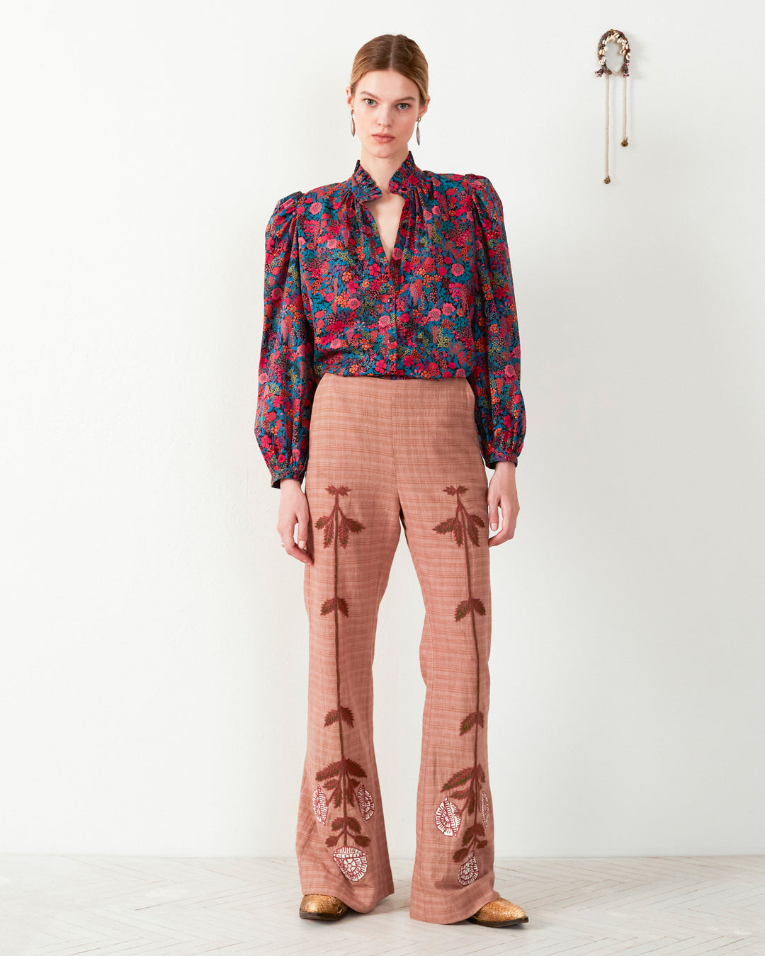 A person stands against a plain wall wearing an artisan's hand-embroidered floral motif blouse paired with Charlie Jute Embroidered Pant. A small decorative item adorns the wall, adding a touch of charm.