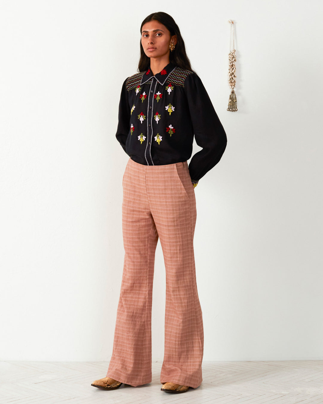 A person stands against a white wall, wearing a black blouse with floral embroidery and the Charlie Juted Rose Pant featuring a hand-stitched hem.