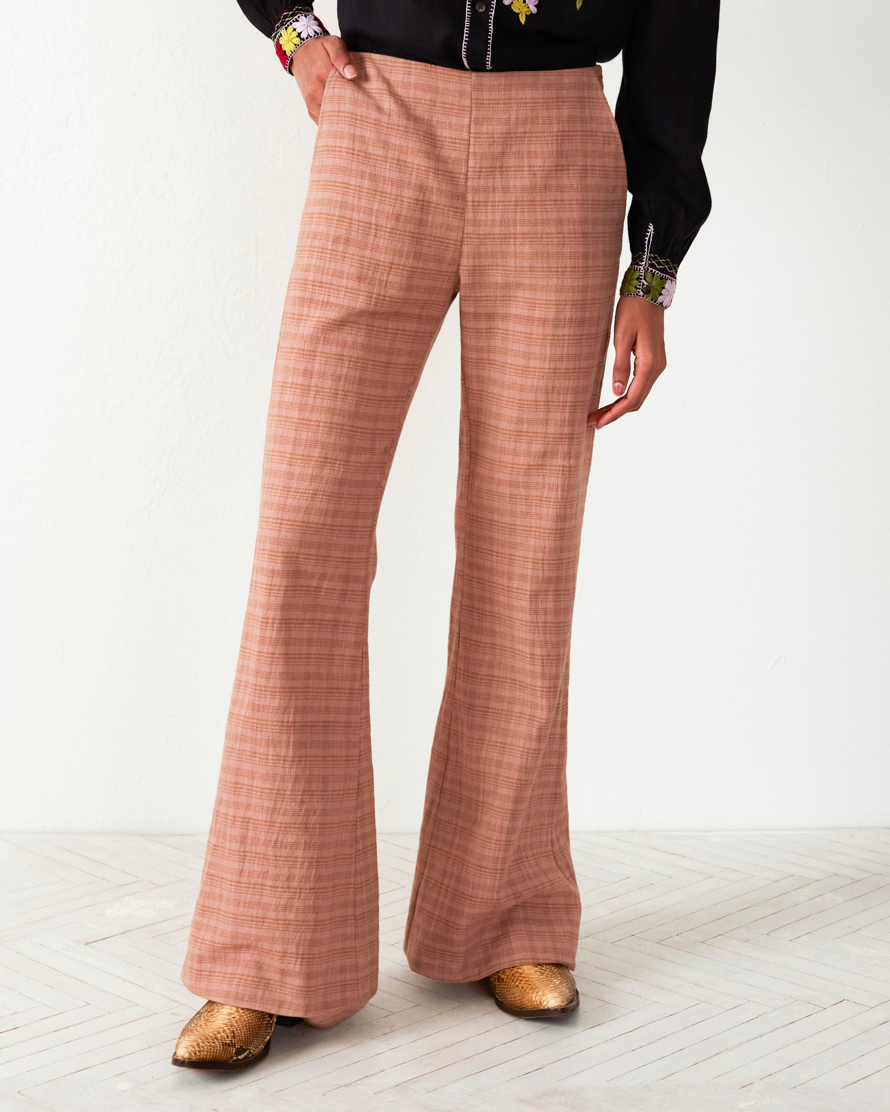 A person wears Charlie Juted Rose Pant in tea rose plaid, paired with a black blouse featuring hand-stitched embroidery on the cuffs. Gold shoes complete the outfit against a light, textured wall and floor.