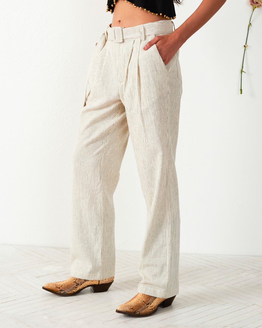 A person dressed in the Colette Cloud Stripe Belted Pants, a black top, and brown cowboy boots stands against a plain background with a single flower on the wall.