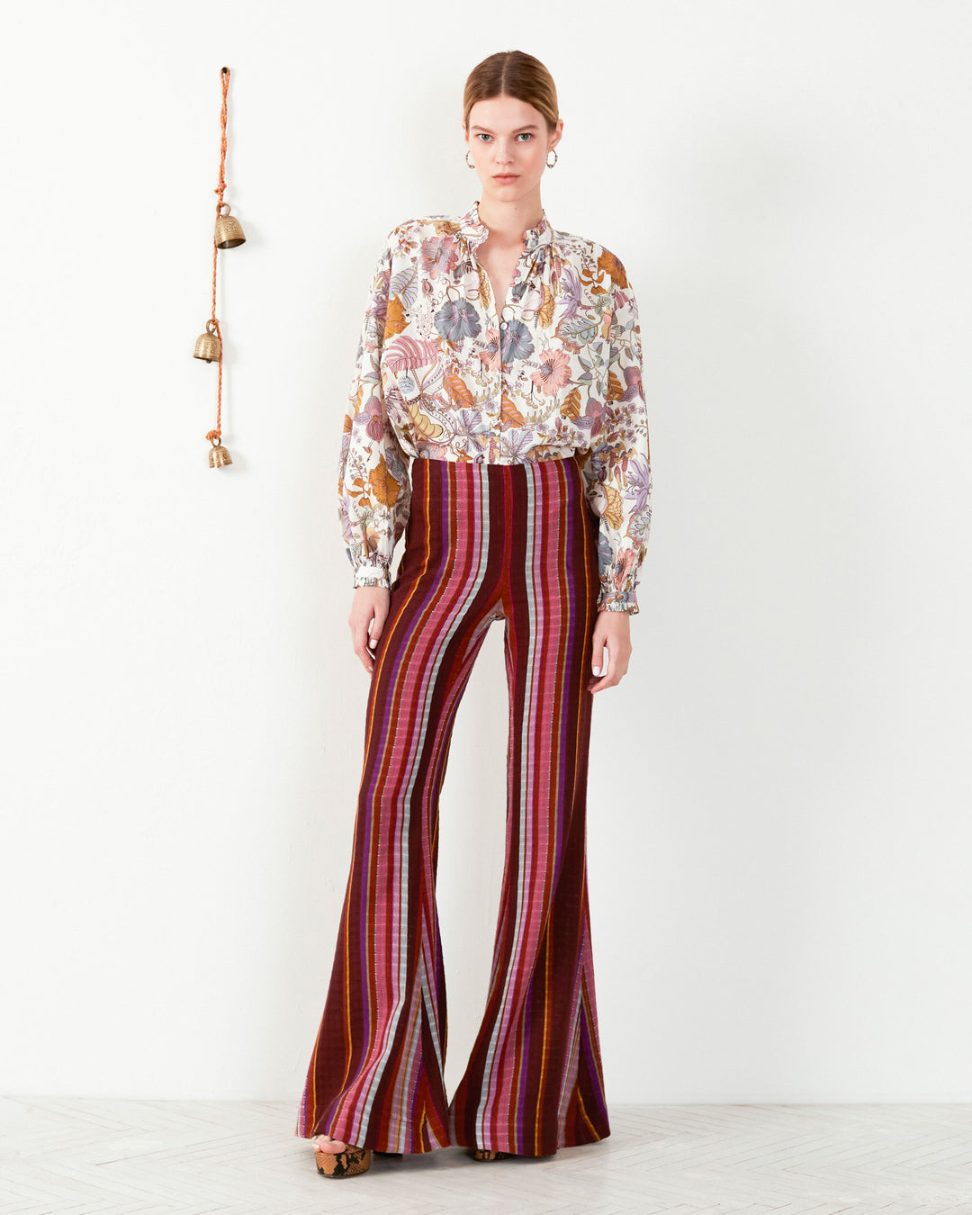 A person stands against a white wall, wearing a floral blouse and the vibrant Farah Berry Truffle Flare Pants. Decorative bells hang on the wall, accentuating this ensemble's mix of patterns and colors for a bold, stylish look with Bohemian flair.