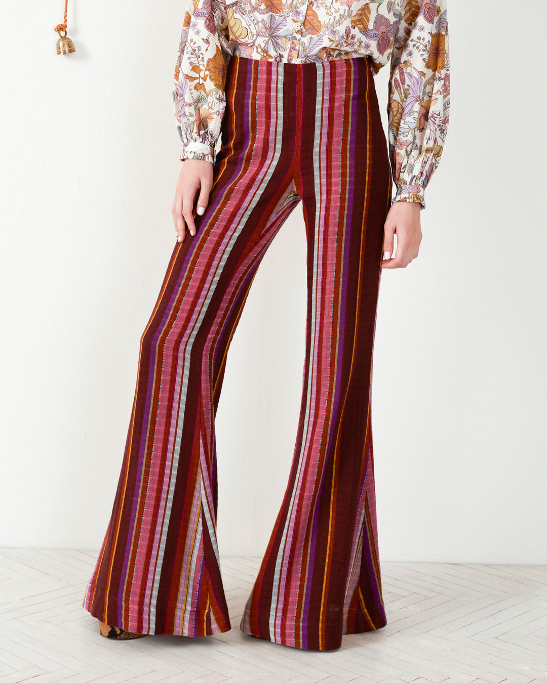 A person is wearing the Farah Berry Truffle Flare Pant, which is high-waisted and features a bohemian style with vertical stripes in shades of red, pink, and purple. The top of the outfit boasts a colorful floral pattern with long sleeves. The floor highlights a light herringbone pattern.