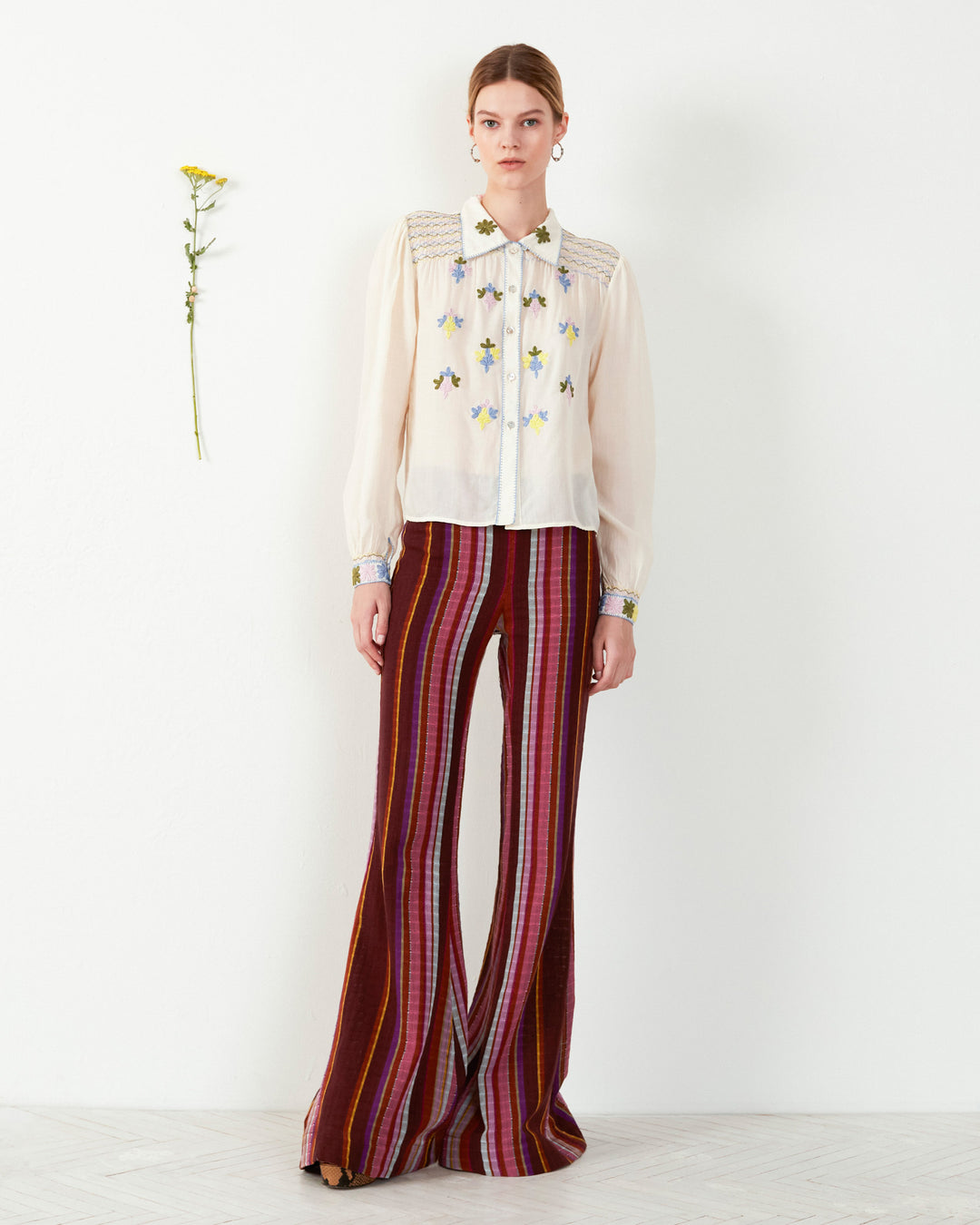 A person stands against a white background wearing a white blouse with floral embroidery and Farah Berry Truffle Flare Pants. A single yellow flower is pinned on the wall beside them.