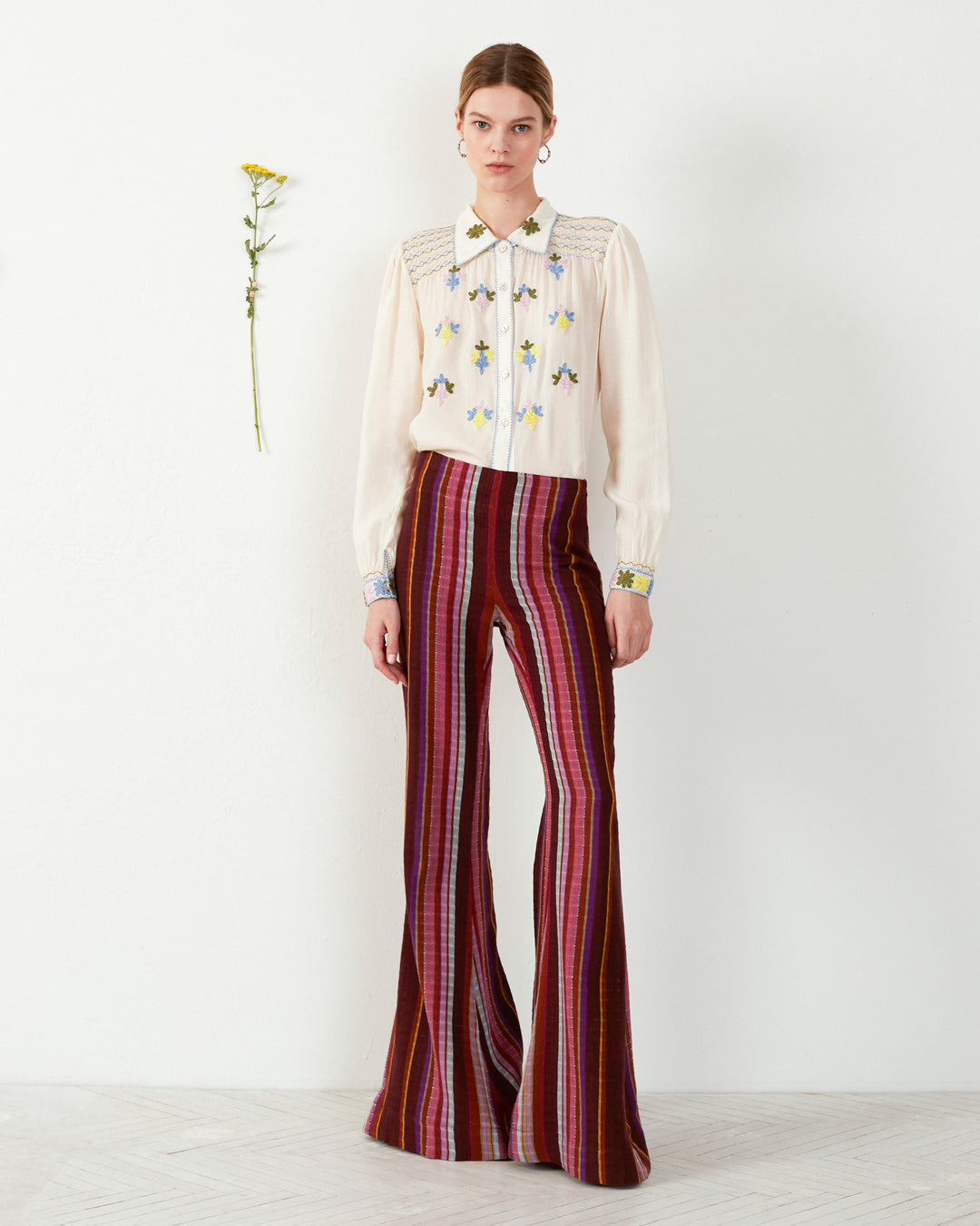 A person stands against a white wall wearing a cream blouse with floral embroidery and the Farah Berry Truffle Flare Pant in maroon. A yellow flower is pinned to the wall beside them.