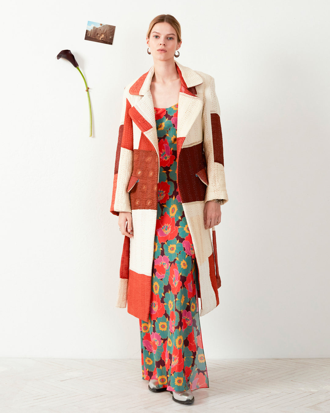 A person stands against a white wall, wearing a colorful floral dress and the Officer Almond Kantha Coat. A single flower and a small photo are pinned to the wall, adding charm to the scene.