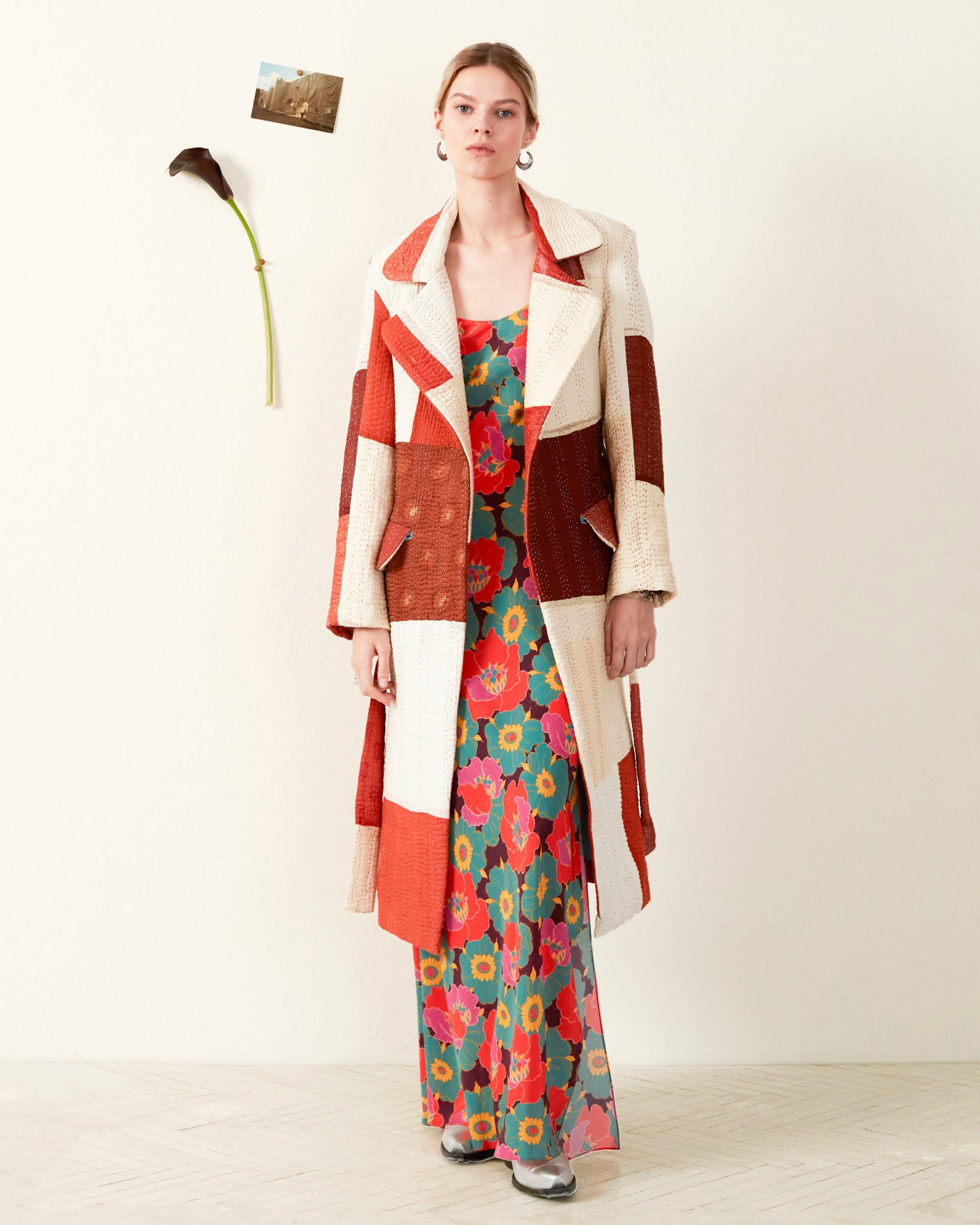 A person stands against a plain wall, wearing the vibrant Officer Almond Kantha Coat made from recycled silk, layered over a floral maxi dress and silver shoes, with a photo and single flower pinned to the wall beside them.