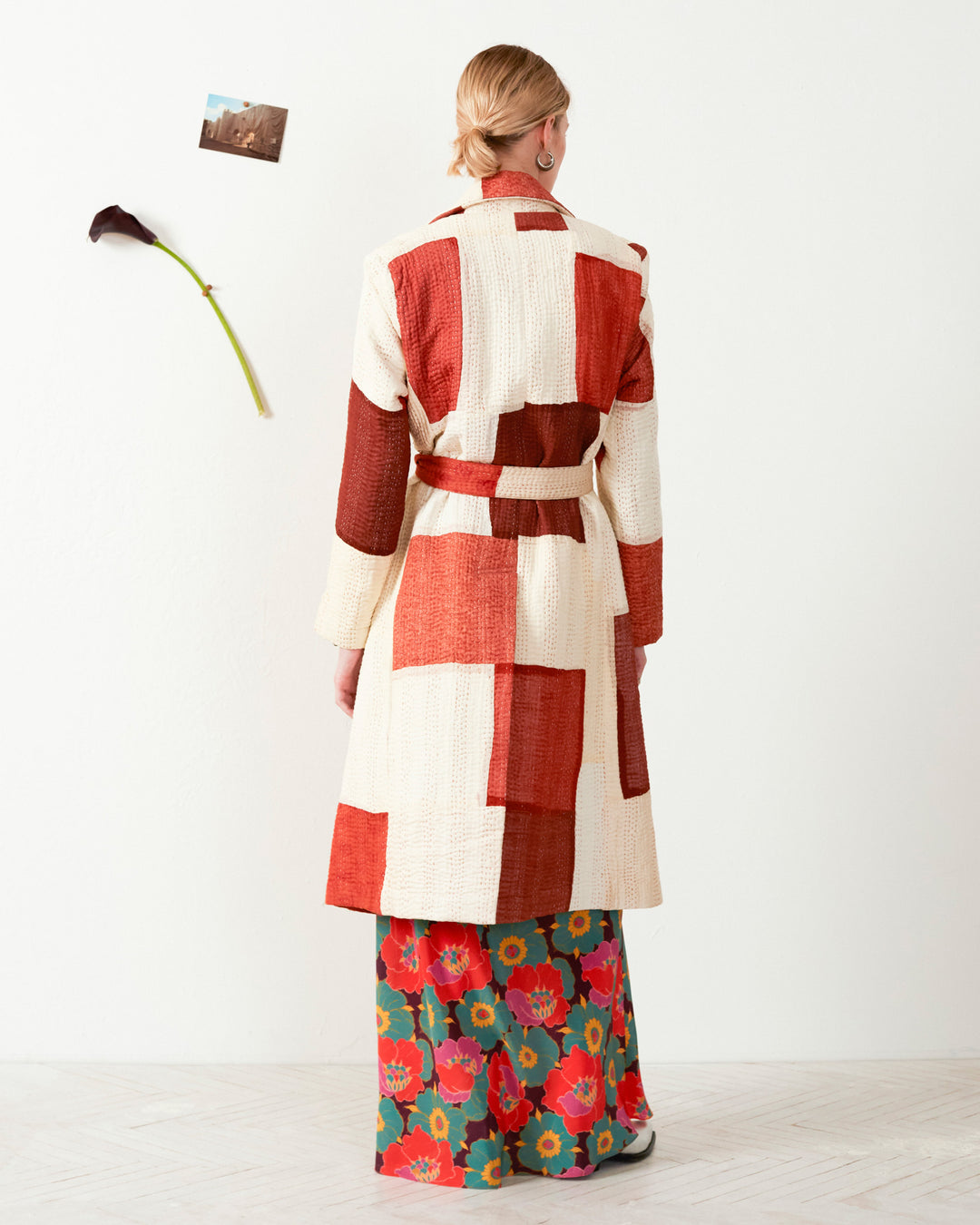 A person wearing the Officer Almond Kantha Coat, featuring red and white quilting, and floral pants stands facing a wall adorned with a flower and photo, highlighting the artistry of hand-stitched craftsmanship.