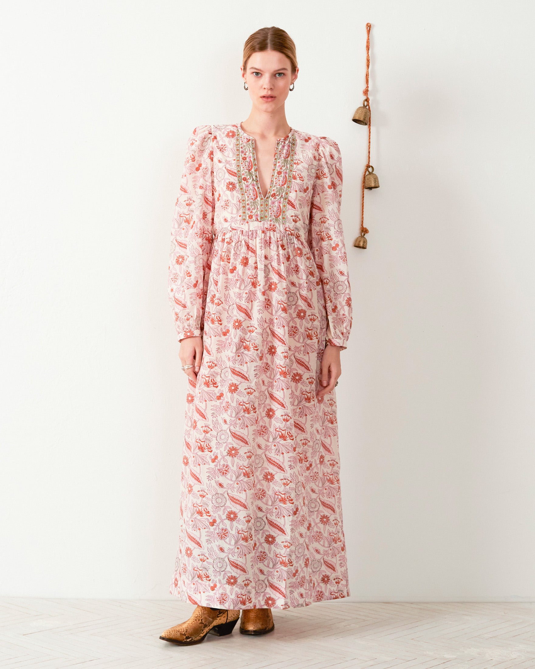 A person stands against a white wall wearing the Winifred Camellia Dress, a floral print maxi dress featuring a deep V-neck, long sleeves, and intricate embroidery on the front. Brown leather shoes complement the ensemble, and a string of bells hangs beside them.