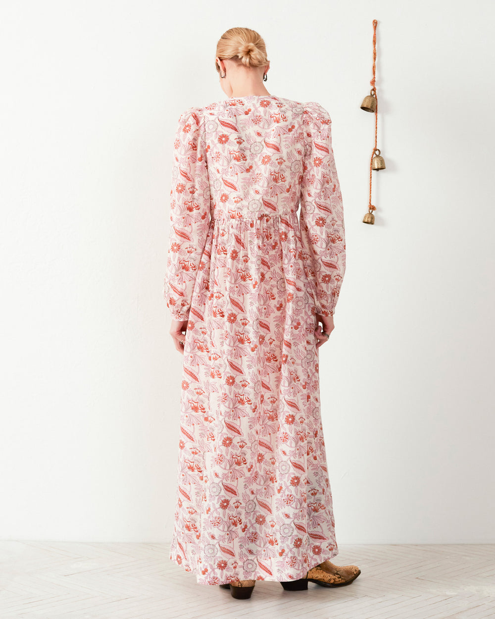 A person stands with their back to the camera, wearing the Winifred Camellia Dress, a long maxi dress with a floral print and long sleeves. The dress is embellished with red and pink tones. They have short hair and are in a minimalistic room featuring a small wall decoration.