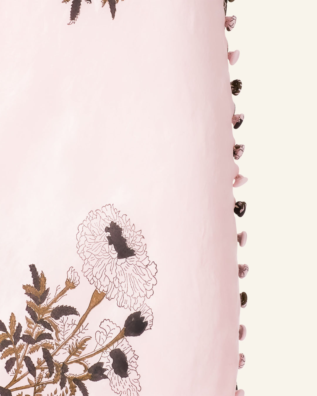 A close-up of the Adele Anemone Silk Dress showcases its light pink block-printed silk, adorned with black and brown floral designs. The edges are enhanced with delicate pink and black tassels, contributing to the fabric's elegant appeal.