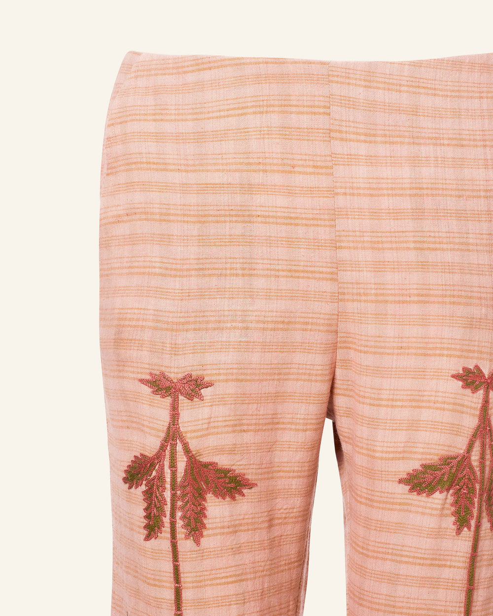 The Charlie Jute Embroidered Pant showcases a close-up of pink plaid trousers with a floral motif, featuring two hand-embroidered designs near the front pockets. The fabric displays a striped pattern, complemented by embroidery of green leaves with maroon accents.