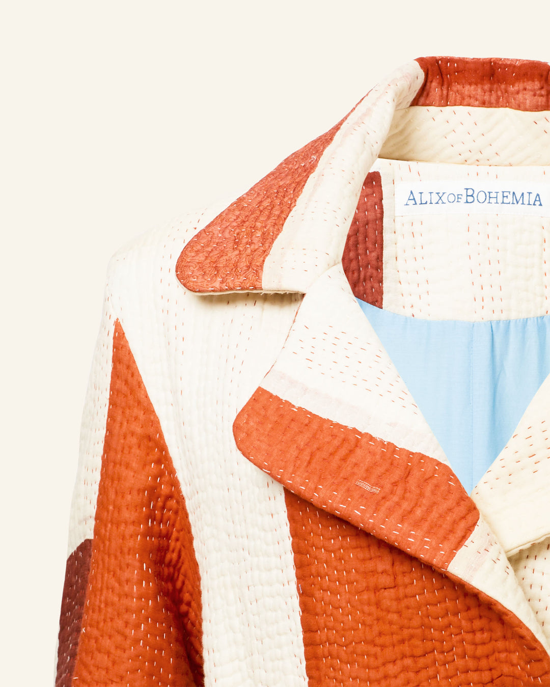 Close-up of the Officer Almond Kantha Coat, a geometric patterned quilted coat in red and cream hues, crafted from recycled silk. Featuring exquisite hand-stitched detailing and labeled as "Alix of Bohemia.