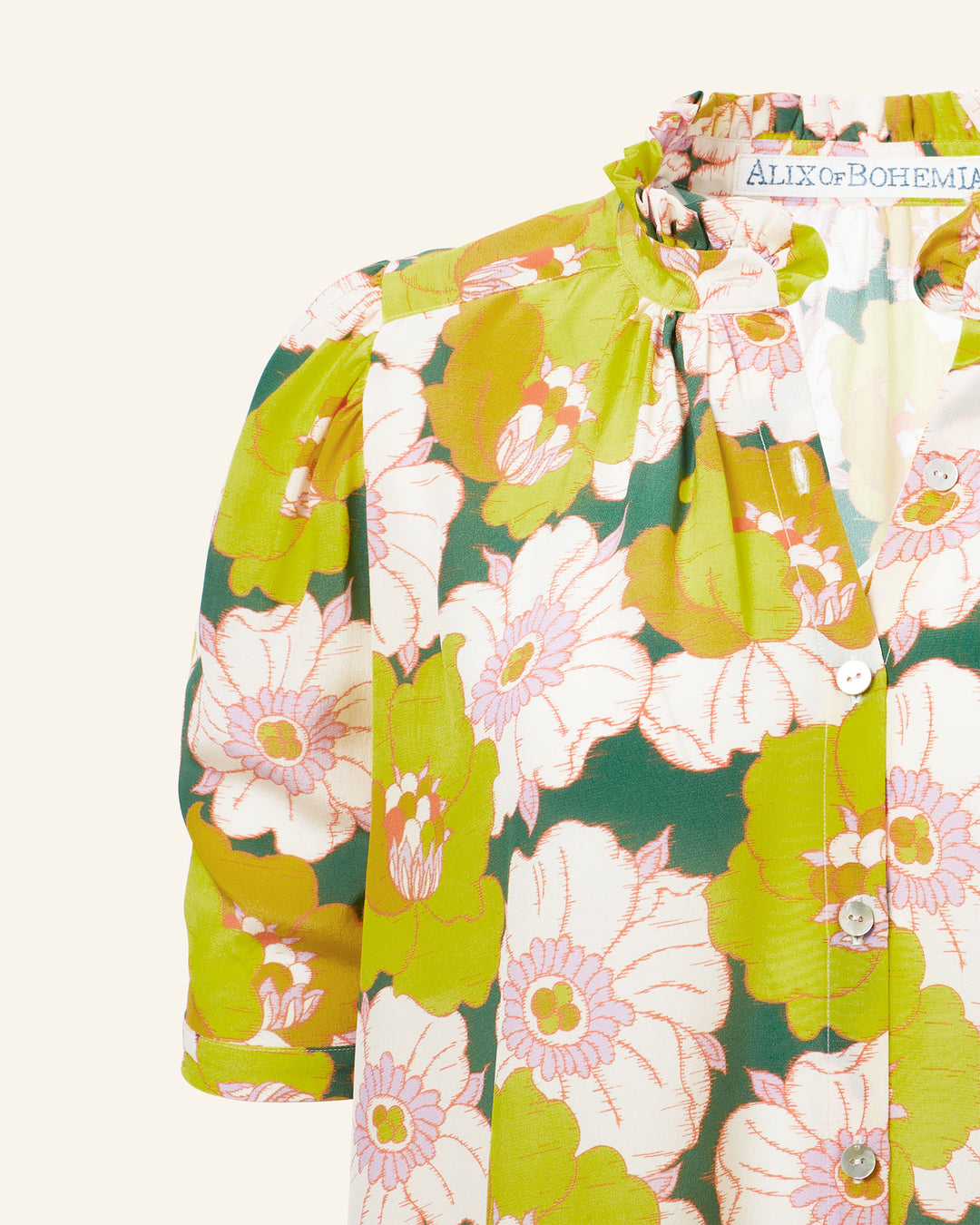 Close-up of the Winnie Citrus Blossom Silk Shirt, crafted from luxurious Italian silk featuring a Liberty-print with green, yellow, and pink flowers. It boasts a ruffled neckline and short sleeves, blending elegance with playful style.