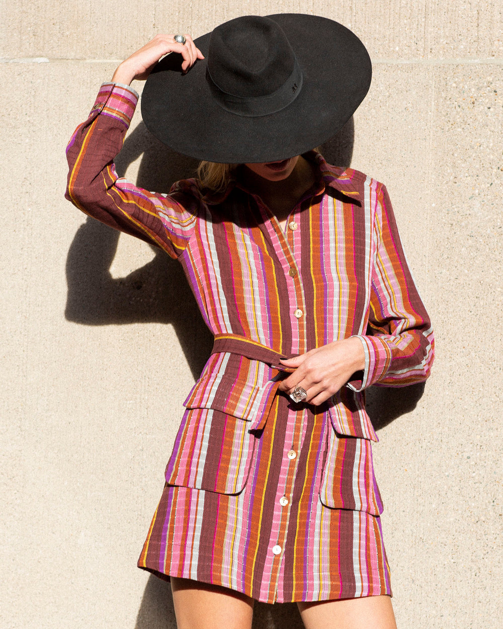 A person wearing a wide-brimmed black hat and the vibrant Arlo Truffle Berry Dress stands against a textured beige wall. The hand-loomed dress showcases a collared neckline and front pockets, with shades of pink, red, and orange. The person's face is partially shaded.