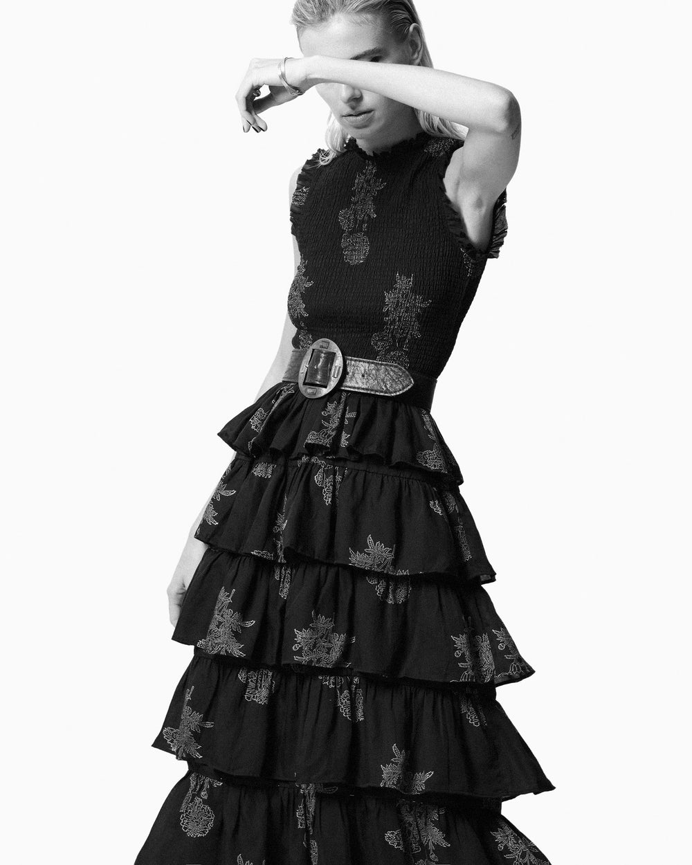 A person wearing the Cabana Ebony Marigold Dress, characterized by its sleeveless black design with a floral print and ruffled skirt, complements their look with a wide belt and covers their eyes with one arm against a plain background. The dress's smocked bodice adds texture to the elegant ensemble.