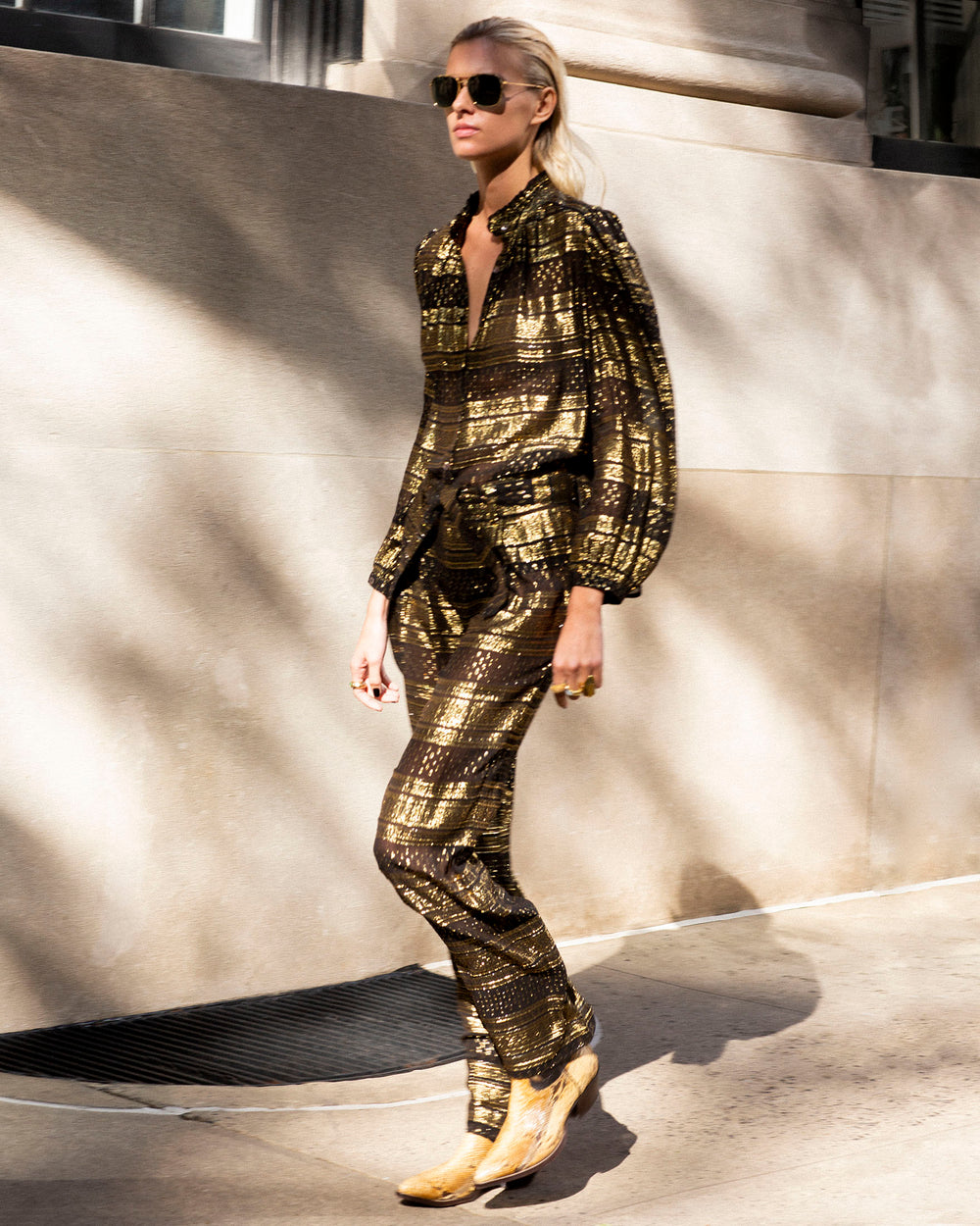 A person dressed in a chic outfit featuring the Colette Gilded Lily Pant, which showcases a gold-check pattern and metallic gold shine, strides along a sunlit city sidewalk. They accessorize with sunglasses and cowboy boots, set against the backdrop of a stone building shadowed by nearby trees.