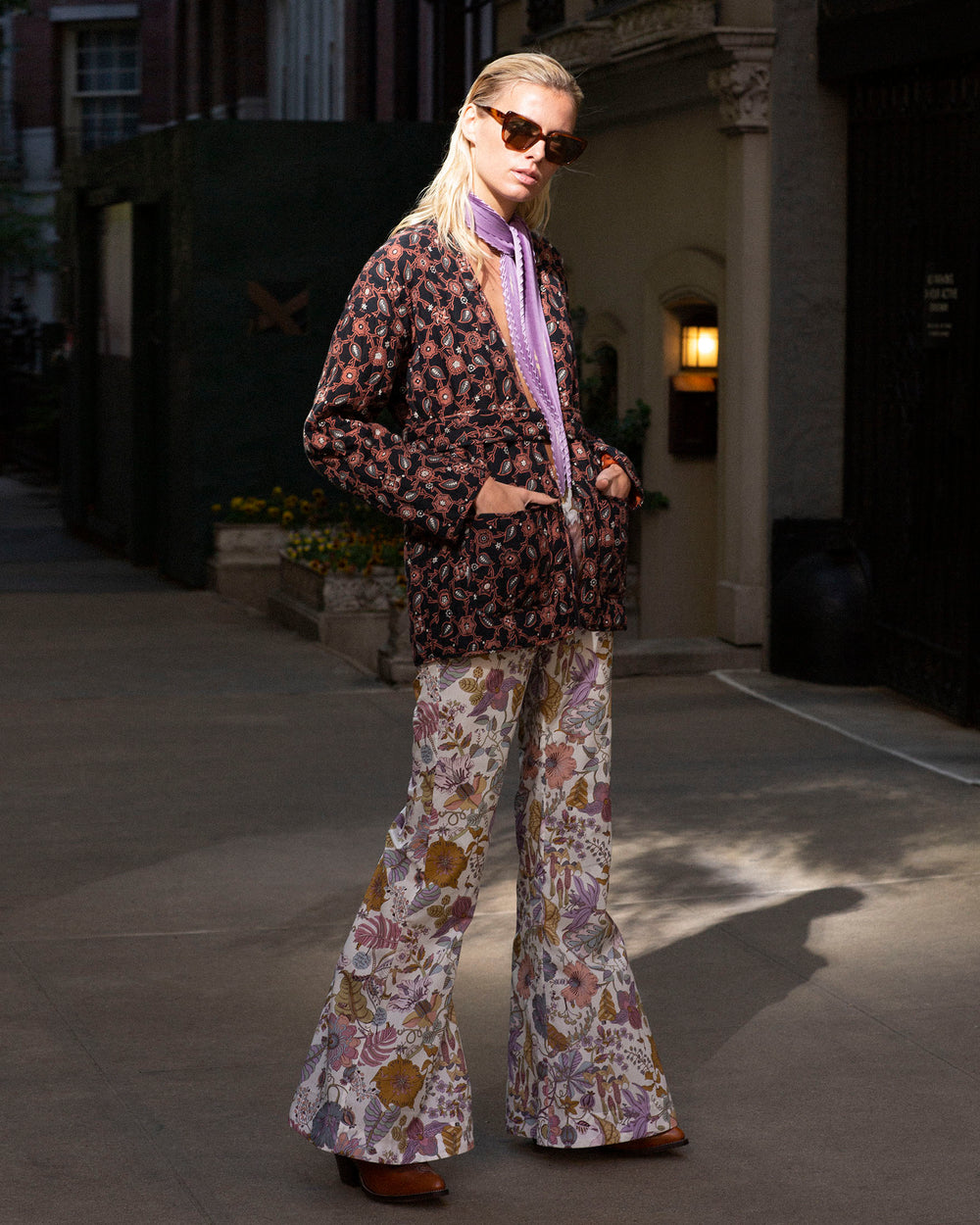 A person wearing sunglasses, a patterned jacket, Farah Faded Eden Flare Pants made of Italian cotton featuring Liberty-print florals, and a purple scarf stands confidently on a dimly lit street. Their attire combines bold prints with a vintage-inspired '70s style vibe, casting a long shadow on the pavement.