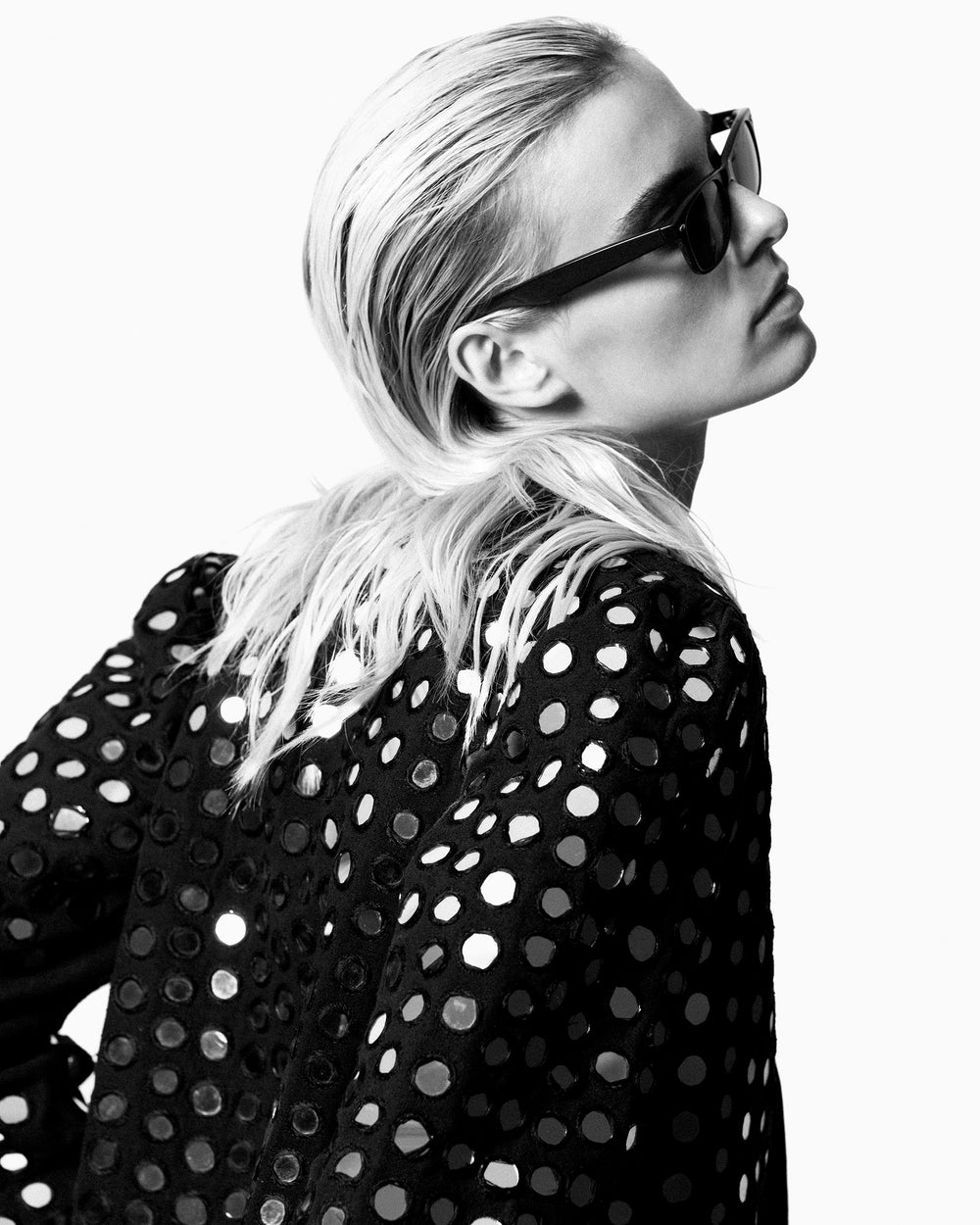 A person with slicked-back blond hair, hidden behind dark sunglasses, dons the Pasha Disco Jacket featuring circular cutout patterns. The black and white image captures their side profile against a plain background, adding an air of mystery and intrigue.