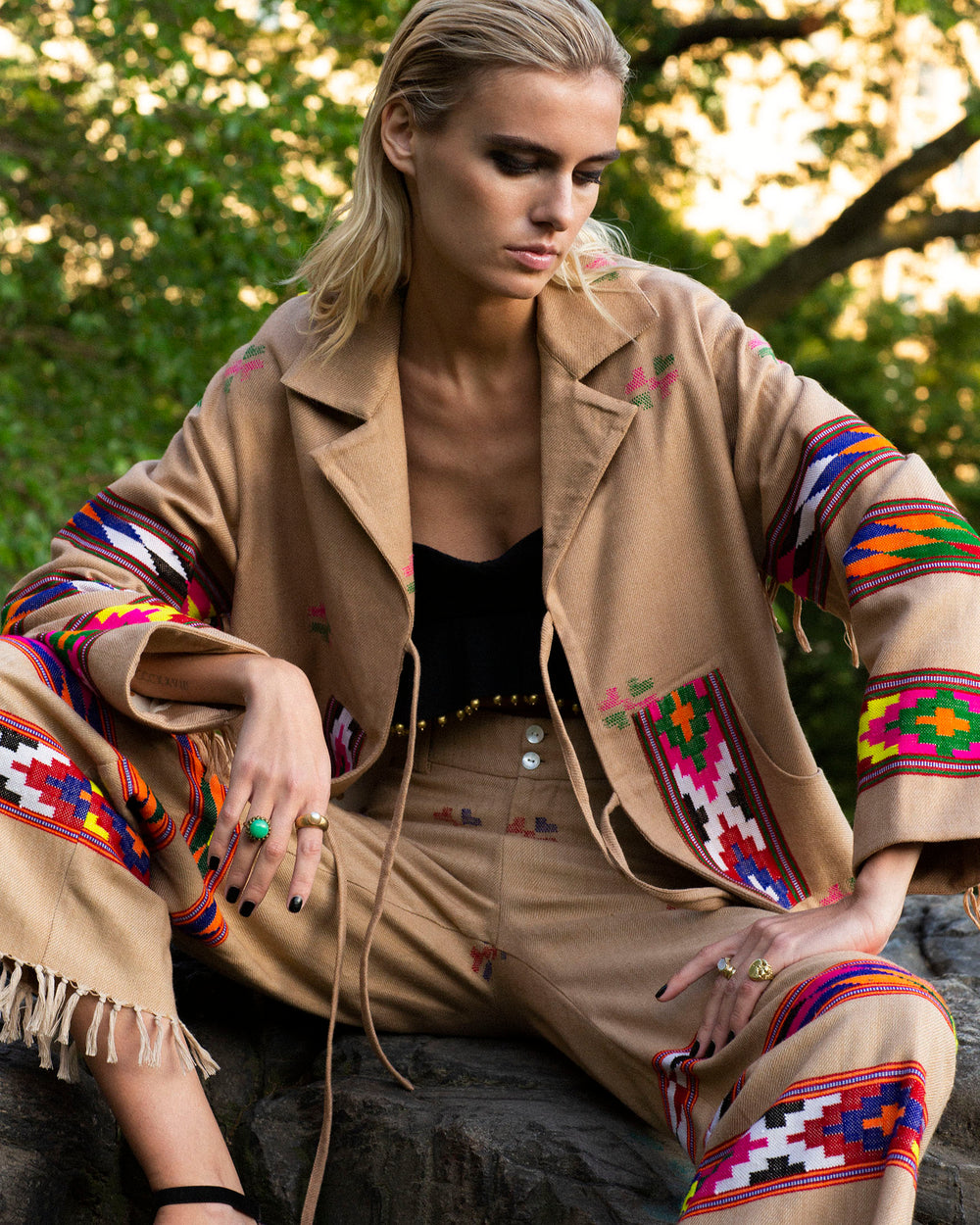 A woman sits outdoors wearing a Western Dune Blanket Jacket made of brown Kullu wool, paired with pants featuring colorful geometric patterns. She has blonde hair and wears black sandals, set against a backdrop of lush green trees.