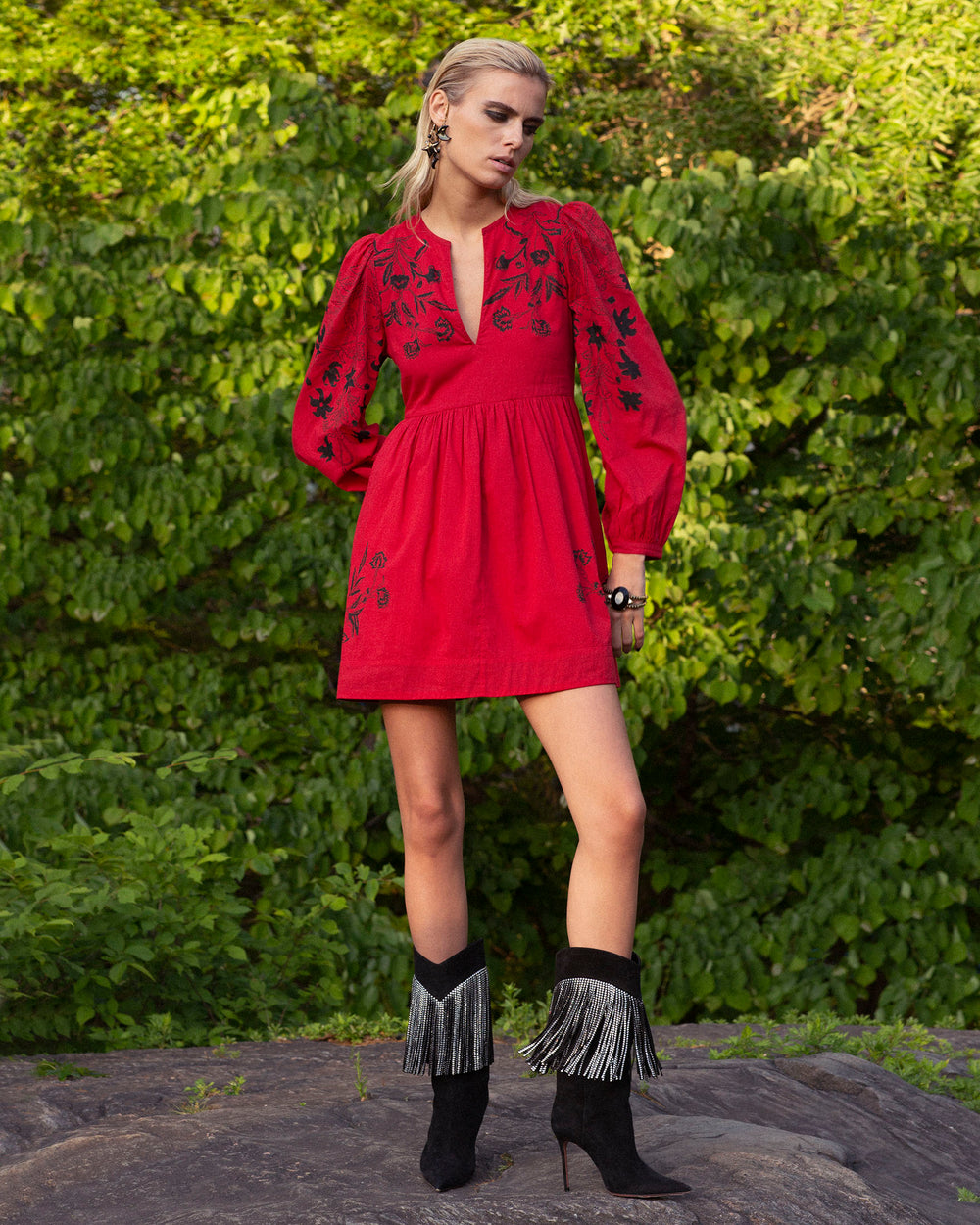 A person stands on a rock wearing the Winslow Cherry Lily Valley Dress paired with black fringe boots, perfectly accentuating the lush green background of leaves.