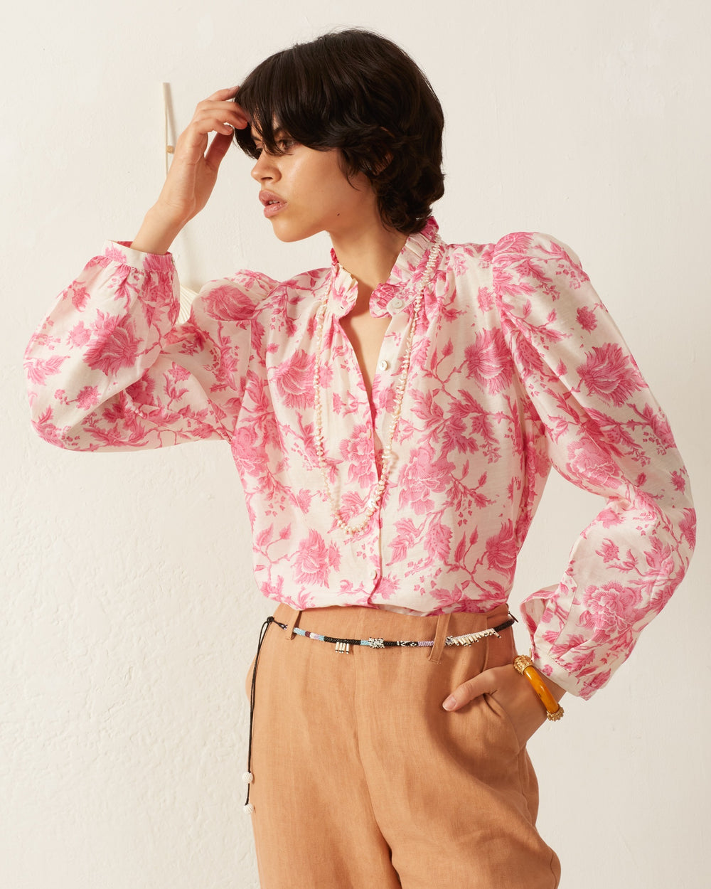 An individual wearing the Annabel Baroque Beam Shirt, a vintage-inspired floral print blouse in pink with puffed sleeves and a ruffled neckline, stands against a white background. They accessorize with beige pants, a thin black belt, and a yellow bracelet, while posing with one hand on their head.