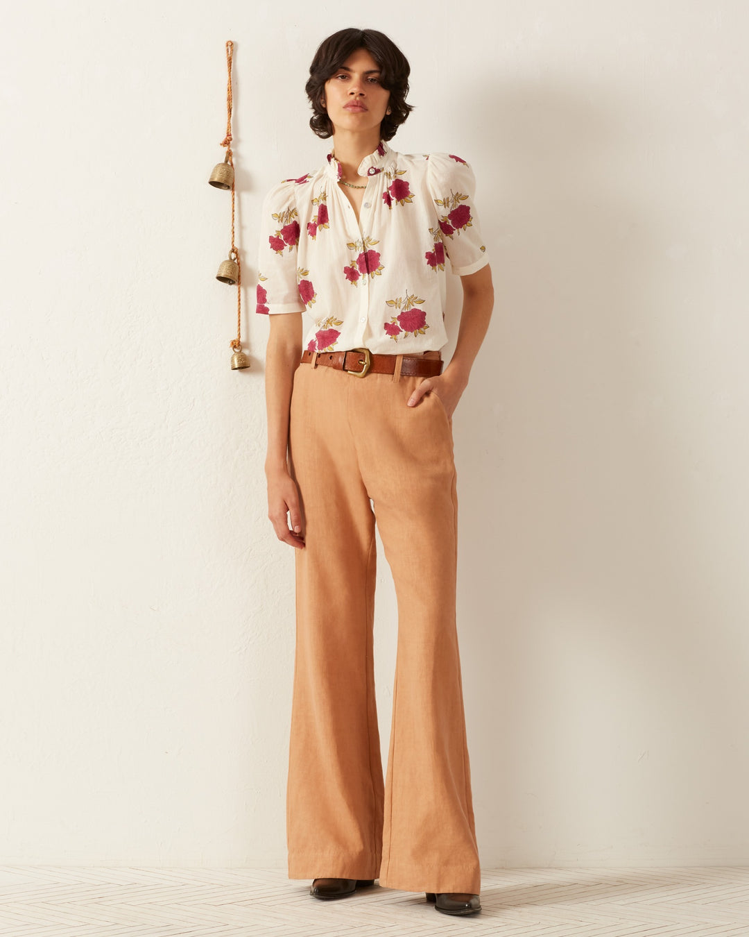 A person stands against a white wall wearing the Winnie Cerise Rosette Shirt, featuring hand-block printed rose motifs and red floral patterns, paired with tan wide-legged trousers. Their short, wavy hair is accompanied by a brown belt and black shoes. A string of bells hangs on the wall nearby, adding a touch of whimsy to the scene.