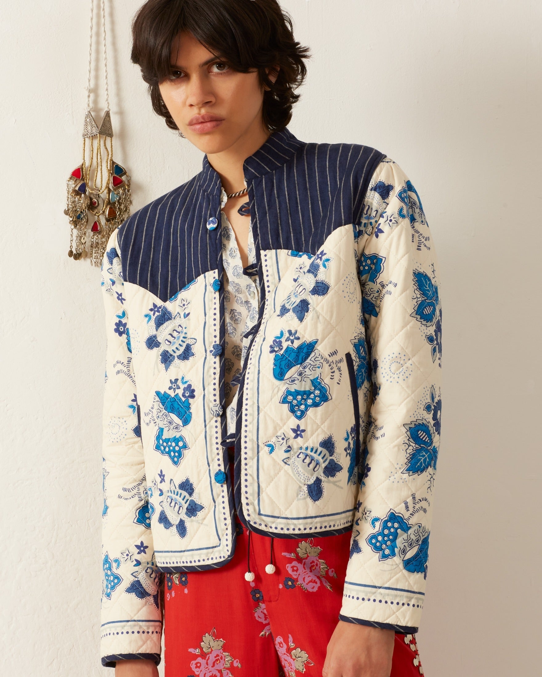 A person with shoulder-length hair wears the Claude Blue China Panel Jacket, which showcases quilted blue floral patterns and a striped yoke. They stand against a neutral background decorated with a hanging piece reminiscent of vintage Provençal textiles.