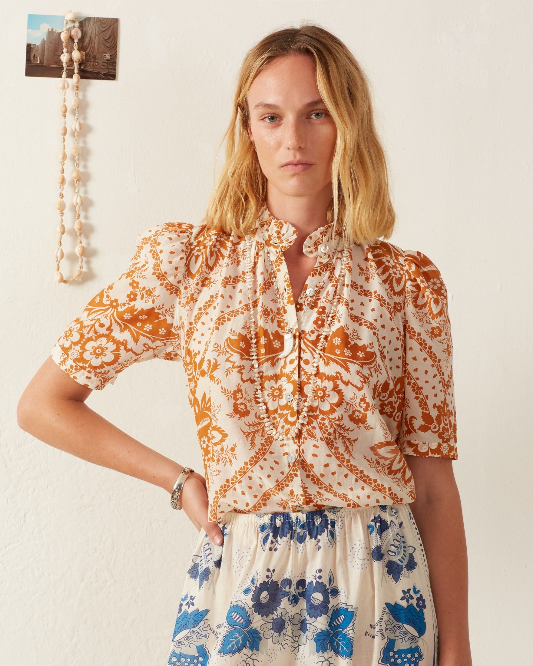 A woman with long blonde hair stands against a cream wall, wearing the Winnie Sunglow Shirt and a matching skirt featuring orange and blue vintage-inspired patterns. The mother-of-pearl button front enhances her elegant appearance. Her expression is neutral, as a small photo and beaded necklace adorn the wall behind her.