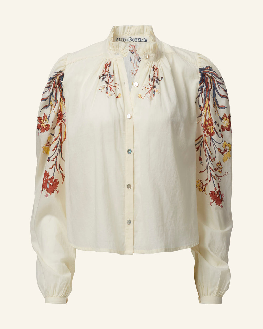 The Annabel Sun Lily Valley Shirt is a cream-colored, cotton voile blouse featuring long sleeves and a button-down front. It is adorned with a floral motif, showcasing intricate embroidery in shades of red, orange, and brown on the shoulders and upper sleeves. This delicate touch enhances the beauty of this hand block-printed garment.