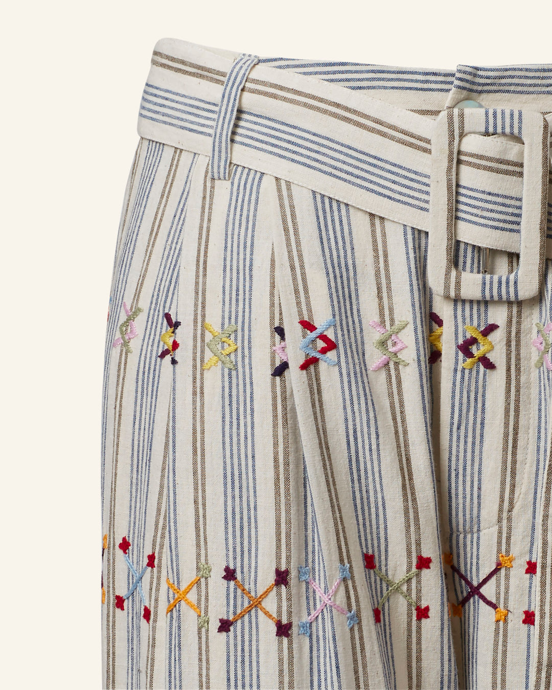 Close-up of the Colette Arrow Linen Pant in khadi cotton, featuring beige fabric with vertical blue and brown stripes. This hand-embroidered piece showcases colorful cross-stitch geometric patterns and includes a fabric belt with a rectangular buckle at the waistband.